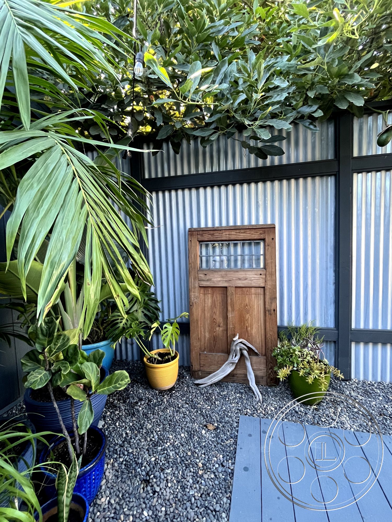 Craftsman 56 - modern rustic design, gravel patio flooring, outdoor garden setting, exterior landscaping features, corrugated metal wall panels, wooden door with glass window, blue painted wooden deck, green foliage accent, potted plants in blue and yellow containers, private outdoor nook, natural texture contrasts, architectural interest with mixed materials