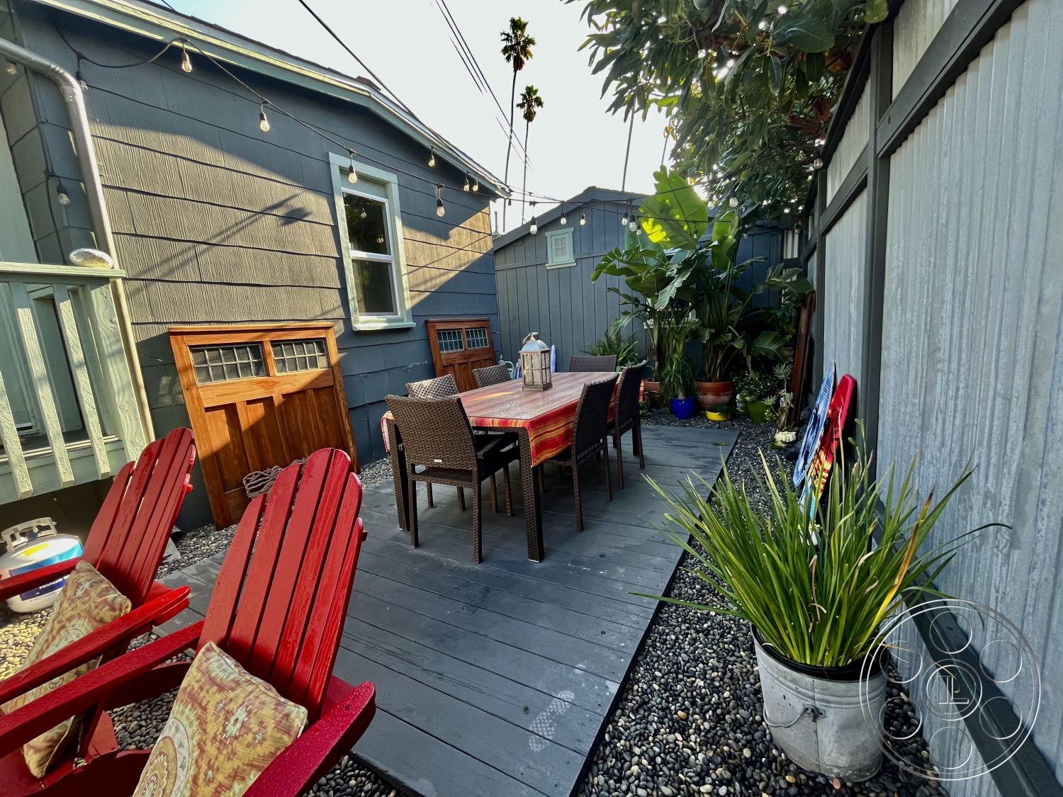 Craftsman 56 - modern outdoor space, outdoor dining table, wood deck patio, potted tropical plants, intimate garden setting, string lights overhead, woven rattan dining chairs, red adirondack chairs, exterior gray shiplap siding, landscape planting beds, pebble garden edging, metal lanterns on table, wood storage bench, interior outdoor dining area, urban patio oasis