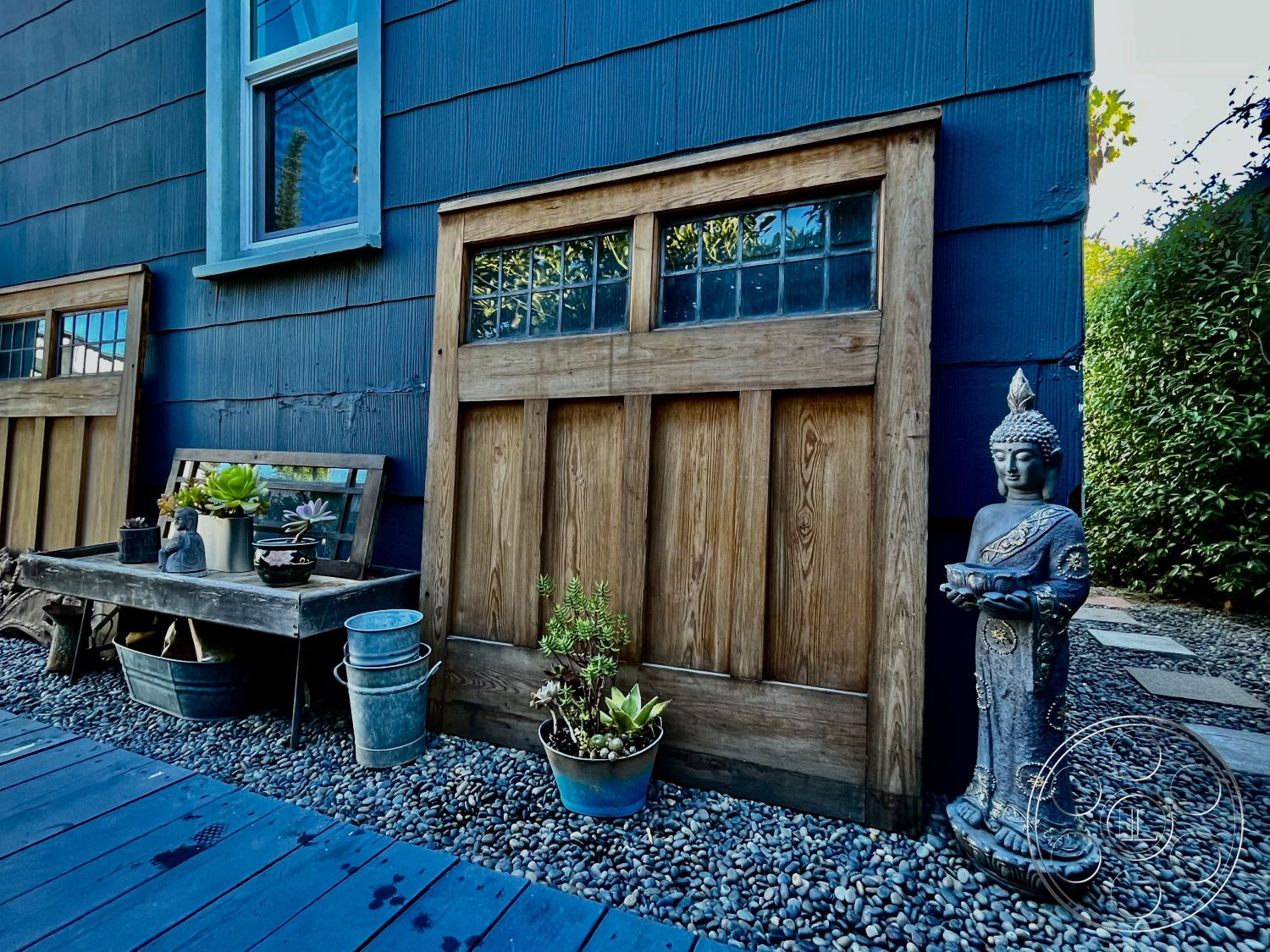 Craftsman 56 - suburban backyard, landscaped garden setting, rustic outdoor decor, wooden garden bench, exterior wood paneling, decorative stone statue, potted succulent plants, natural stone stepping stones, exterior blue painted walls, exterior wooden window frames, exterior double-hung windows, exterior wooden door panels, outdoor gravel pathway, metal garden buckets, architectural Craftsman style details