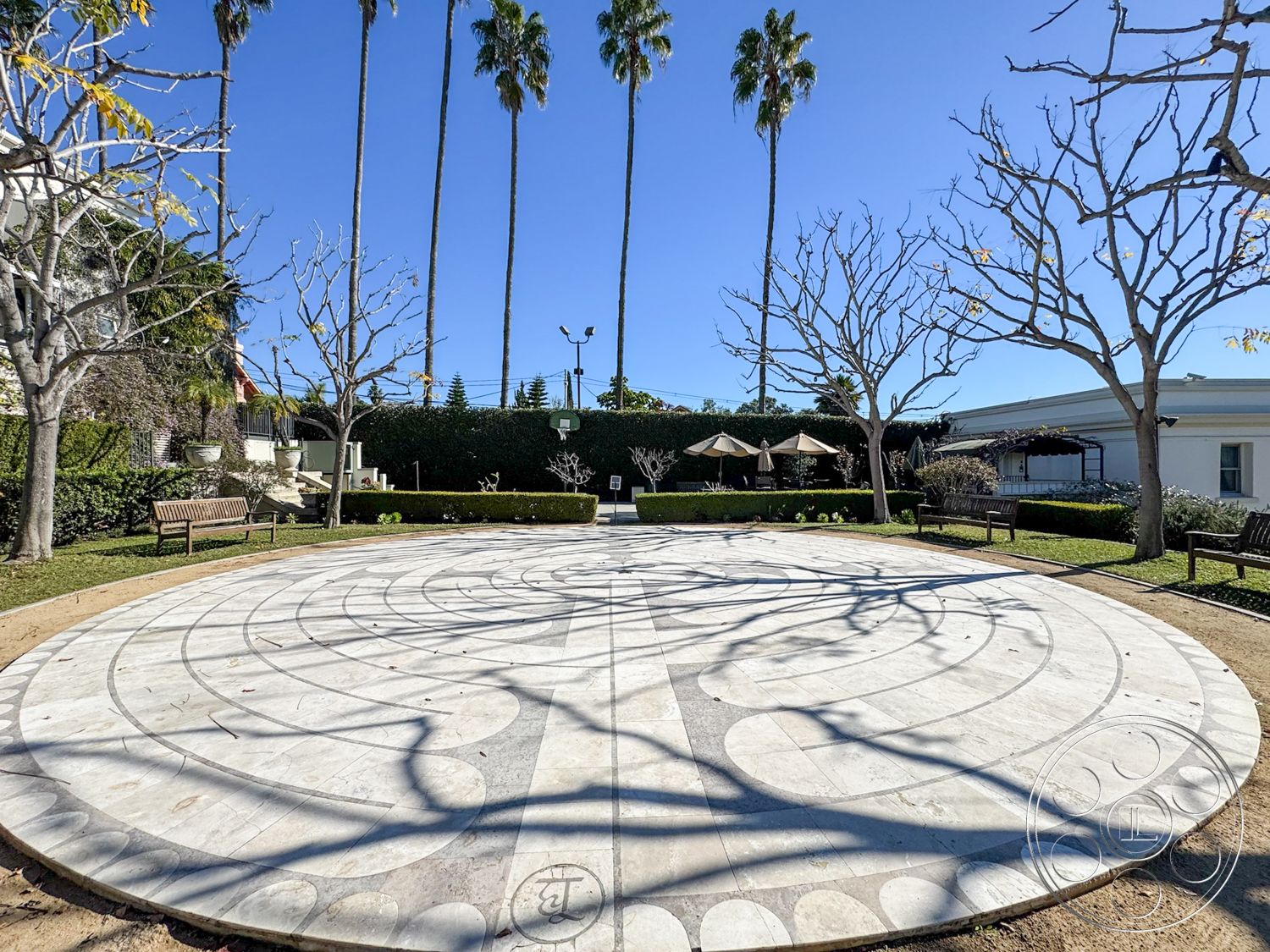Adams Mansion - exterior garden area, landscape with palm trees, landscaped lawn area, benches for seating, tranquil outdoor setting, circular marble patio flooring, geometric pattern marble design, decorative low hedge borders, sunken garden environment, textured concrete walkways, seasonal tree planting, shadow patterns from overhead trees, outdoor recreational area, urban garden destination