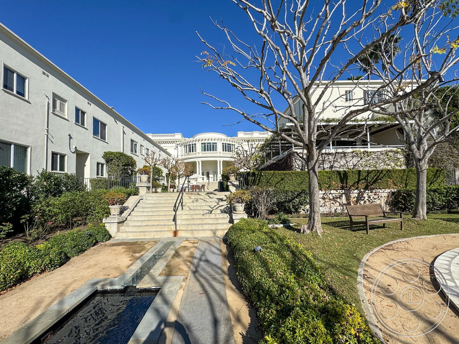 Adams Mansion - floor-to-ceiling windows, modern two-story home, stucco exterior walls, urban garden setting, landscaped garden with hedges, concrete planter boxes, hardwood bench seating, concrete staircase with railing, stone pathway with water feature, rectangular front patio, exposed tree branches, luminary lighting fixtures, architectural columns leading to entrance, balustrade railings, paved courtyard area, shaded outdoor dining area, formal entrance with steps, surrounding greenery landscape, symmetrical building facade