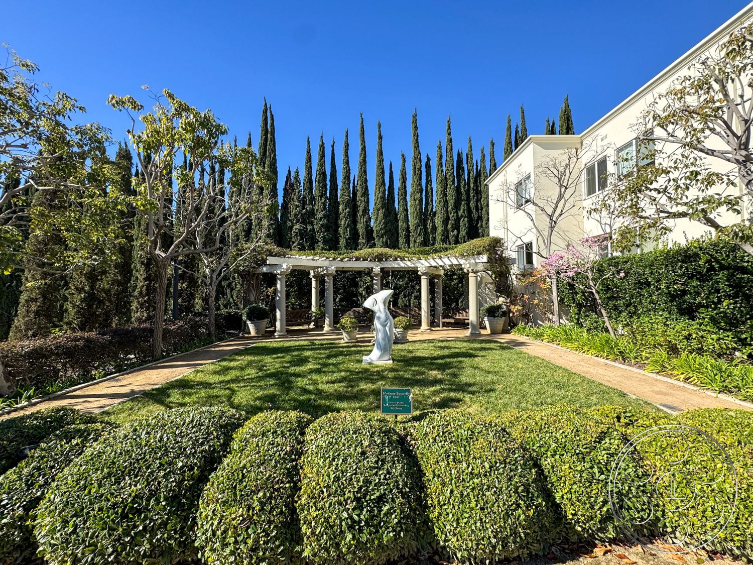 Adams Mansion - exterior garden setting, lawn area with manicured grass, pergola structure with white columns, sculpture in garden, cypress trees providing privacy, hedge landscaping for borders, brick pathway leading through garden, framed garden view with blue sky, neighboring two-story home, flowering shrubs adding color, stone patio floor beneath pergola, arched trellis design, natural sunlight illuminating space, suburban backyard ambiance