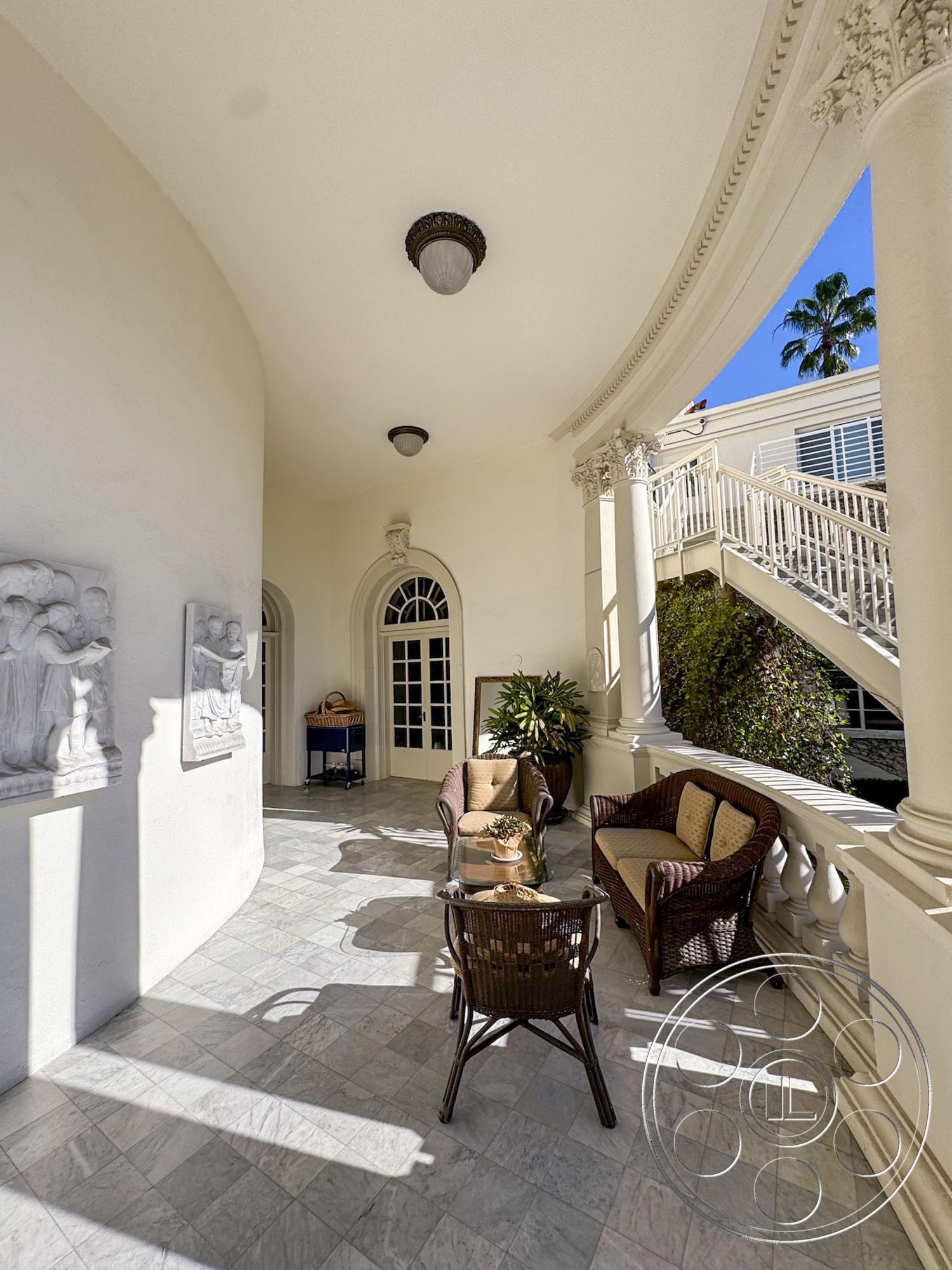 Adams Mansion - colonial architectural style, white textured plaster walls, tropical plant accents, luxurious outdoor living space, exterior marble flooring, arched doorways with elegant details, exposed decorative ceiling moldings, wicker outdoor seating, glass coffee table on patio, detailed stone wall sculptures, curved porch design, lawn view with palm trees