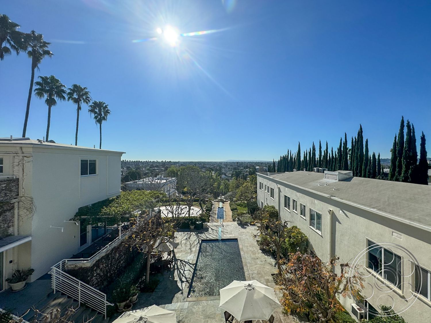 Adams Mansion - exterior, rectangular swimming pool, urban skyline view, concrete pool deck, paved driveways, stone patio flooring, symmetrical building layout, climbing vines on walls, tiled pool border, white stucco building facade, landscape of palm trees, glass patio furniture, terraced garden area, hardscape with gravel pathways, overhead sunshade umbrellas, architectural details with large windows, modern coastal setting, vibrant green landscaping, casual outdoor seating areas
