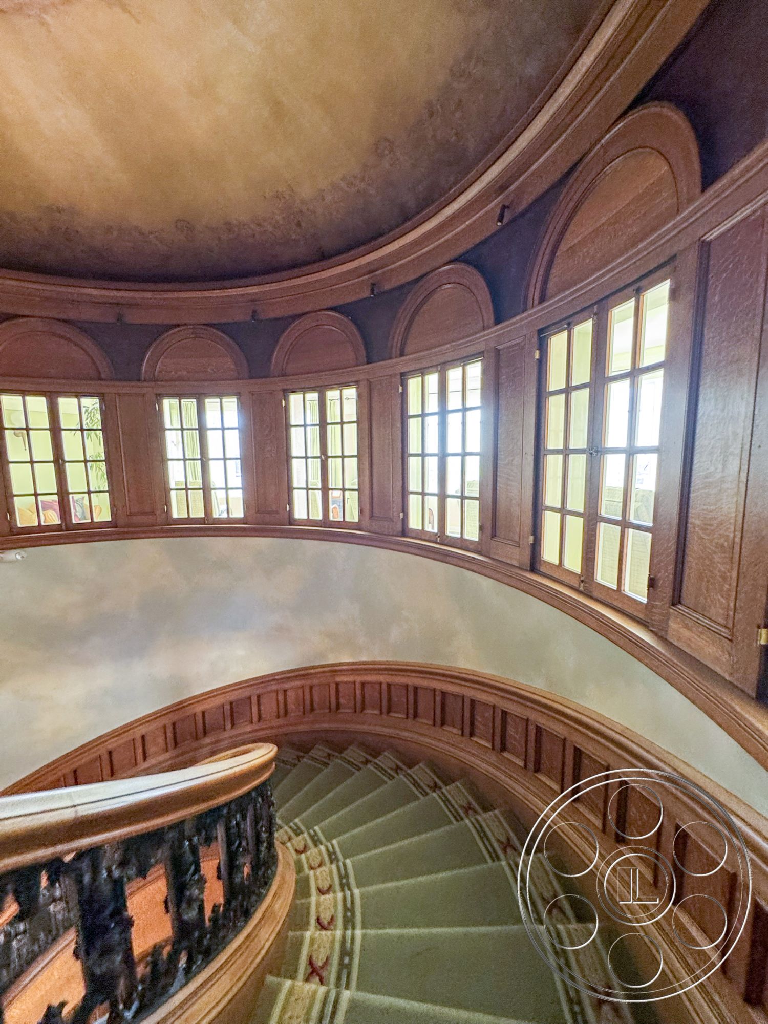 Adams Mansion - natural wood tones, exposed wood ceiling beams, wood paneling accent walls, historical architectural details, interior grand foyer, elegant interior finishes, curved staircase with carpet flooring, arched doorways with natural light, floor-to-ceiling windows with multiple panes, intricate iron stair railing, vaulted ceiling with decorative finishes, ornate window trim in circular layout, textured plaster wall treatment in stairwell, classic Victorian style, soft lighting for stairwell ambiance