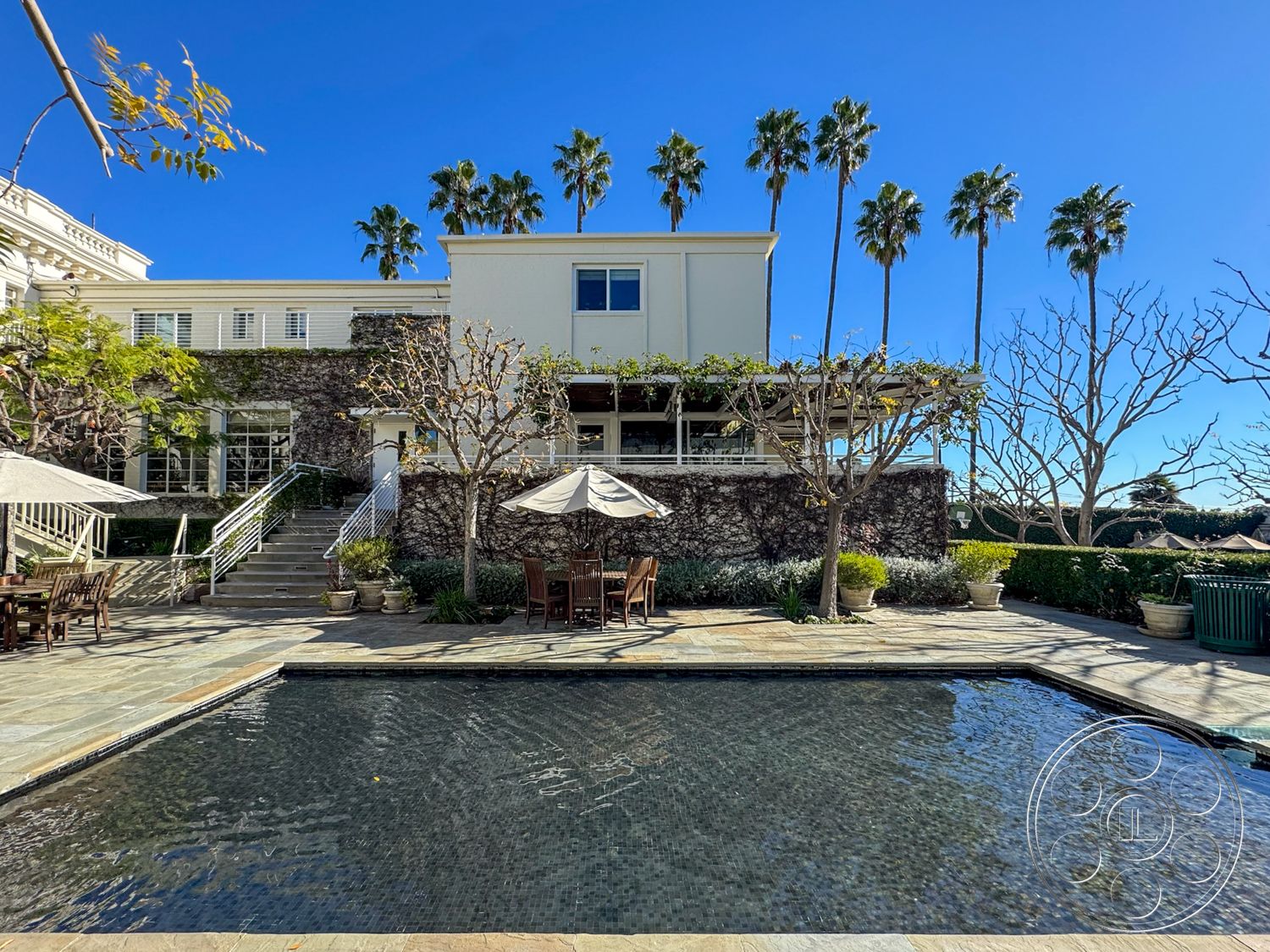 Adams Mansion - exterior, glass sliding doors, stucco exterior, rectangular pool, exposed stone wall, terraced garden, lush green landscaping, stone pool deck, two-story home, patio umbrella, iron railings, landscaped garden area, concrete patio floor, palm tree surroundings, sunny exterior setting, wooden outdoor dining set