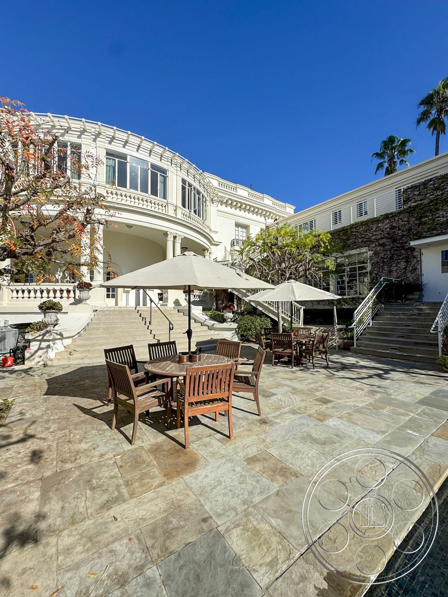 Adams Mansion - glass railing, floor-to-ceiling windows, formal entryway, two-story home, exterior stone patio flooring, lush garden setting, palm tree landscaping, exterior textured stone walls, modern outdoor living space, luxury residential property, exterior sandstone patio tiles, exterior wooden outdoor dining set, classical architectural details, curved stairway, ornate column design, parasol shade structures, elevated terrace area, expansive outdoor seating area, arched entries, sunny coastal view