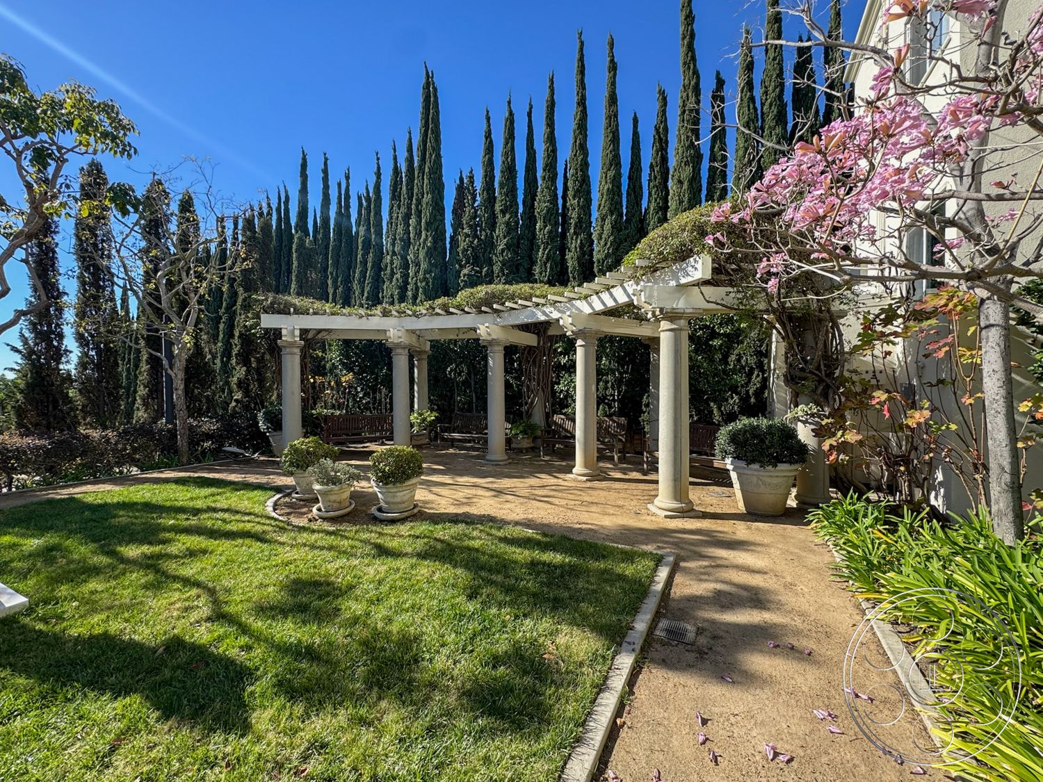 Adams Mansion - outdoor seating area, green grass lawn, stone patio flooring, sunlit garden space, traditional garden setting, exterior pergola structure, planted landscaping beds, tall cypress trees backdrop, flowering cherry tree, concrete path edging, ornamental pot planters, columned support for pergola