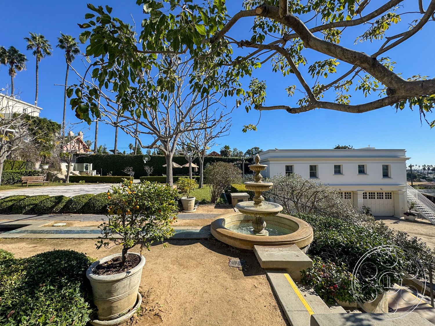 Adams Mansion - outdoor seating area, desert landscape, colorful flowering plants, lush green hedges, elevated outdoor area, circular stone fountain, landscaped garden setting, urban garden space, white stucco building exterior, two-story residential home, exterior fountain feature, paved walkway with stone pavers, hardwood garden bench, tree canopy overhead, rectangular fountain basin, sculptural outdoor feature, potted plants in clay planters, palms in the background, minimalist hardscaping, natural sandy ground cover