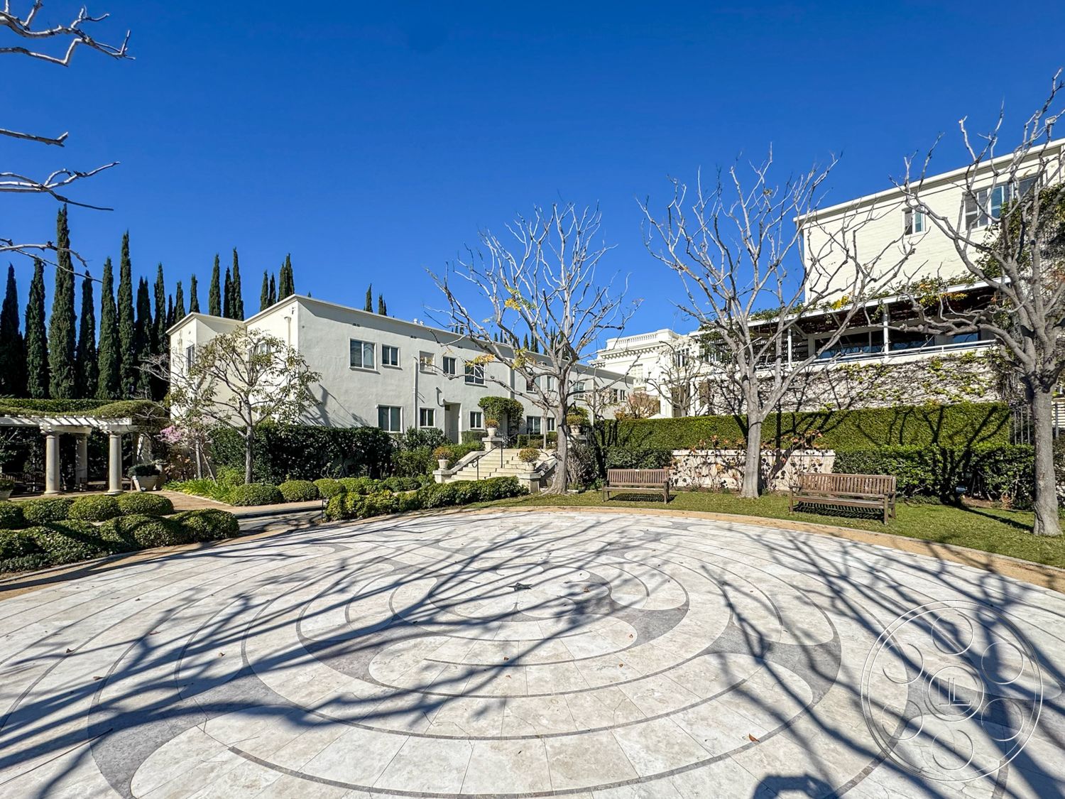 Adams Mansion - exterior, decorative landscaping, smooth concrete pathway, modern two-story home, urban garden setting, stone patio flooring, minimalist architectural style, lush greenery surrounding patio, landscaped garden with hedges, white stucco wall finish, outdoor gathering area, blue sky backdrop, circular stone seating area, pergola with wooden beams, natural stone accent wall, contemporary garden benches, symmetrical tree alignment, geometric circular design