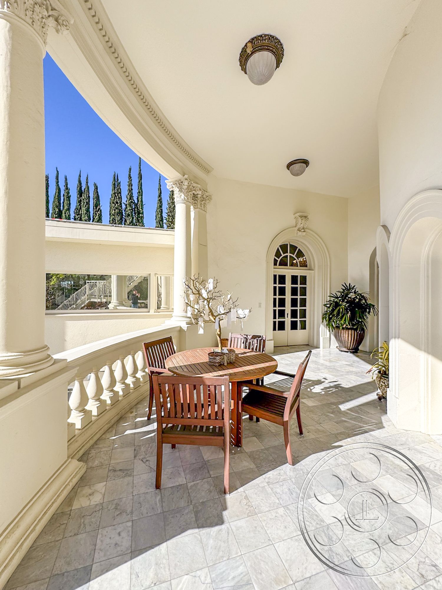 Adams Mansion - exterior marble patio flooring, curved balcony with decorative railing, wood dining table with slatted design, classic plaster wall finish, ornate ceiling light fixture, tall cypress trees in background, arched doorways leading to living spaces, columned architectural details, potted plants for landscaping, natural light from surrounding windows