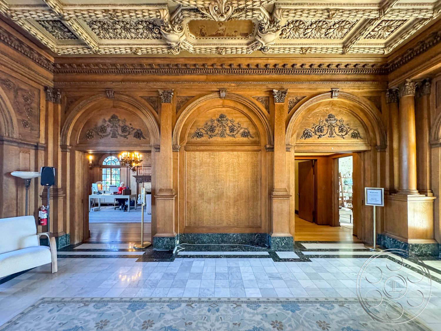 Adams Mansion - interior framed artwork on walls, interior recessed lighting in ceiling, interior coffered ceiling with ornate detailing, interior wooden wainscoting, interior arched doorways with decorative trim, interior marble flooring with intricate patterns, interior dark granite baseboards, interior antique chandelier lighting fixture, interior spacious entry hall design, interior wooden accent walls with carvings, interior polished marble tile floor, interior plush upholstered armchair, interior historical architecture features, interior open layout connecting to adjacent rooms, interior traditional style decor elements, interior contrasting color palette of wood and marble