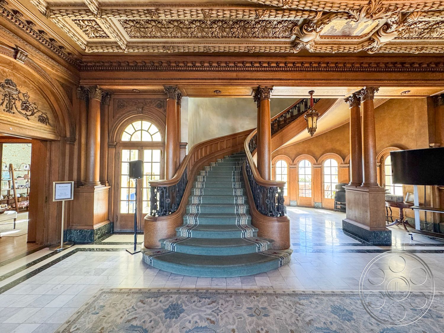 Adams Mansion - interior natural light from large windows, interior soft color palette, interior wood paneling walls, interior vintage area rug, interior decorative plaster details, interior ornate coffered ceiling, interior curved staircase with carpet runner, interior marble flooring with decorative border, interior carved wooden balusters, interior large arched windows, interior chandelier lighting fixture, interior dark wood columns, interior wainscoting detail, interior brass stair railings, interior elegant foyer space, interior grand entrance hall, interior classic architectural features, interior multi-level staircase design, interior historical revival style, interior spacious gathering area