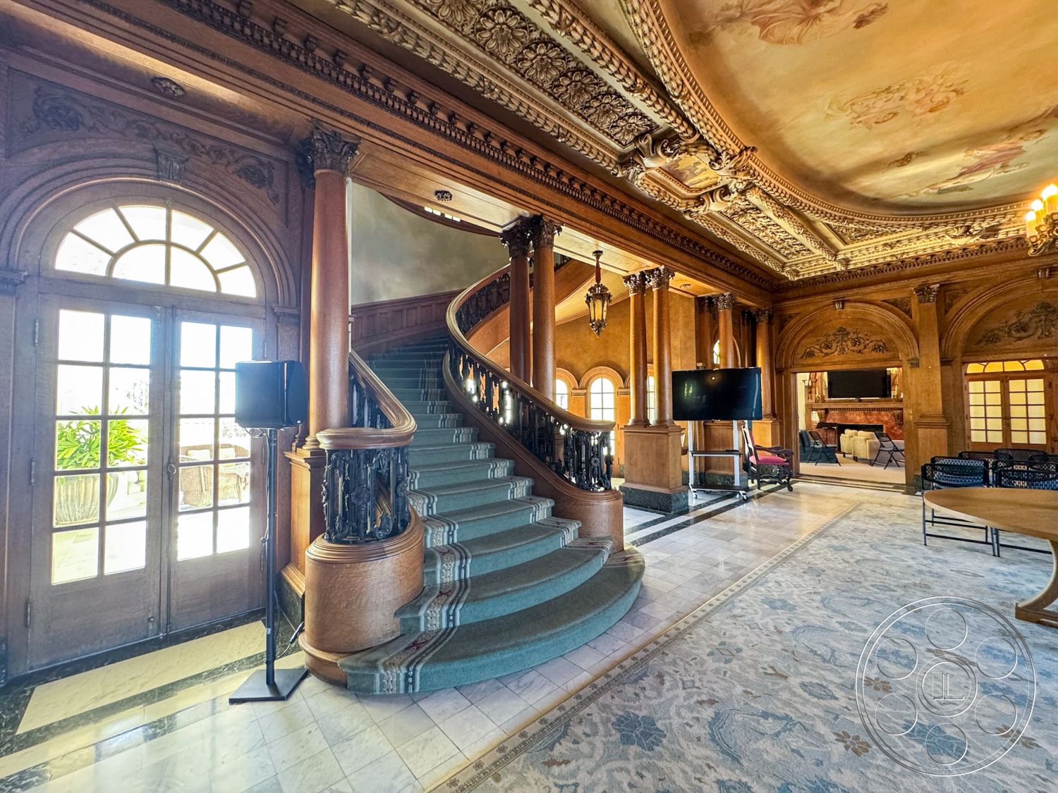 Adams Mansion - large arched windows, textured wall treatments, high vaulted ceilings, open floor plan layout, natural light flooding the space, exposed wooden ceiling beams, dark wood paneling accents, marble entryway flooring, historical architectural style, chandelier lighting fixtures, ornate plaster ceiling details, intricate wood staircase with curved forms, luxurious area rugs with floral designs, built-in decorative shelving, traditional fireplace with stone surround, ornamental crown molding, elegant half-moon entry door, stylish living area with seating arrangements, expansive view of garden patio, rich wood finishes