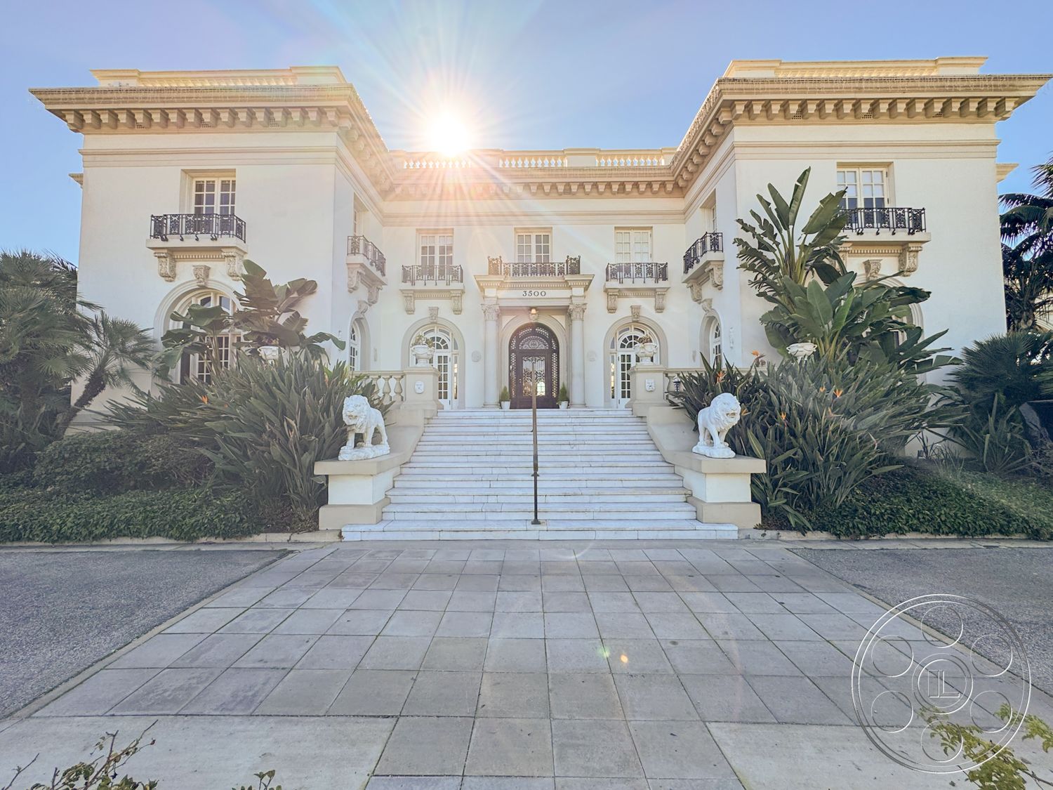 Adams Mansion - high ceilings, exterior, symmetrical facade, ornate ceiling details, white stucco facade, sunlit exterior, landscaped garden with tropical plants, paved stone driveway, classical architectural style, marble entrance steps, ornamental iron balcony railings, arched double entry doors, tall windows with decorative molding, lion statue flanking staircase, detailed cornice molding, header stone lintels, formal front entrance, elegant stair risers, large open courtyard, luxurious residential setting