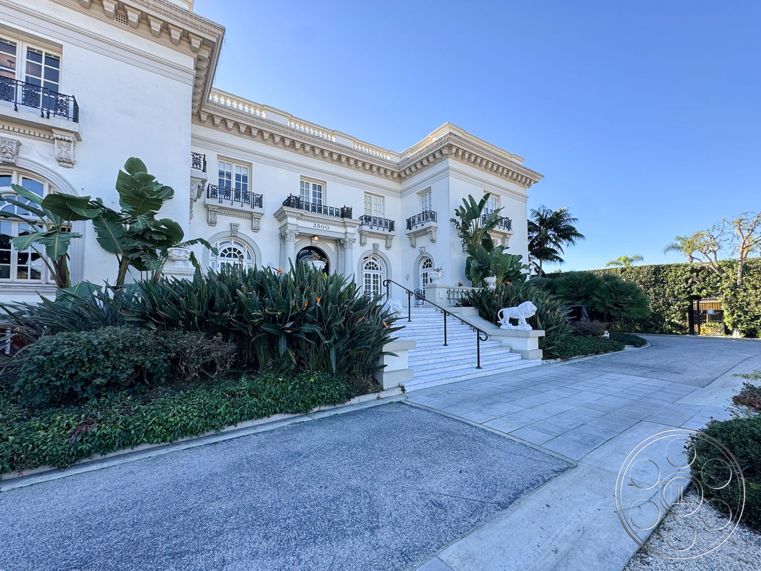 Adams Mansion - exterior, suburban neighborhood, paved pathway, concrete driveway, curved driveway, arched doorways, large multi-pane windows, formal entryway, landscaped garden area, classical column accents, tropical plant landscaping, white painted stucco walls, ornate architectural detailing, marble front steps, black wrought-iron balcony railings, historic mansion style, elaborate exterior facade, luxury residence setting, north-facing entrance, decorative lion statue, private property gates