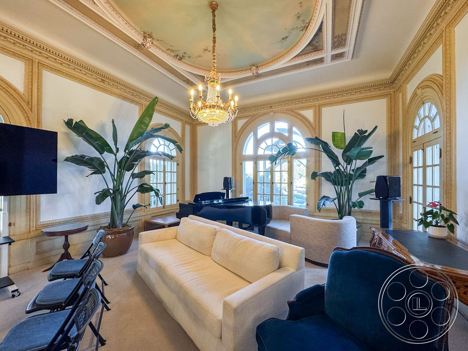 Adams Mansion - black grand piano, large arched windows, natural light from window, interior living room, potted indoor plants, neutral carpet flooring, beige upholstered sofa, dark wood side table, exposed plaster ceiling with decorative molding, gold chandelier lighting fixture, green velvet armchair, trimmed wall details, elegant decorative wall accents, traditional style architecture, spacious and airy room layout