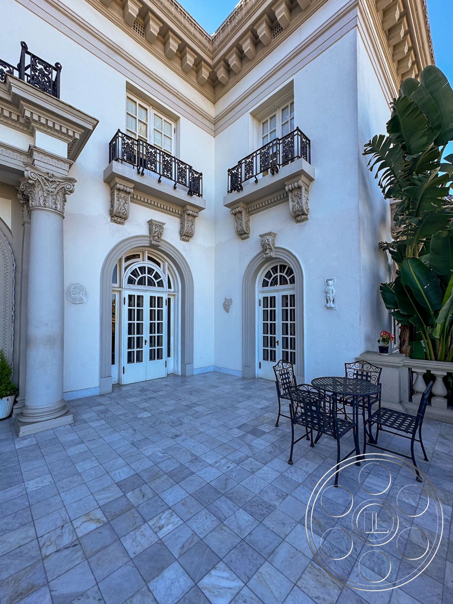 Adams Mansion - Mediterranean architectural style, interior coffered ceiling details, suburban backyard setting, exterior double French doors, interior grand entrance, exterior marble patio flooring, exterior decorative wrought iron railings, exterior classical column supports, exterior ornate window trim, exterior stone planter boxes, exterior traditional balcony design, exterior potted tropical plants, interior white stucco wall finish, interior symmetrical architectural style, interior transitional space, interior courtyard layout, vintage patio furniture set