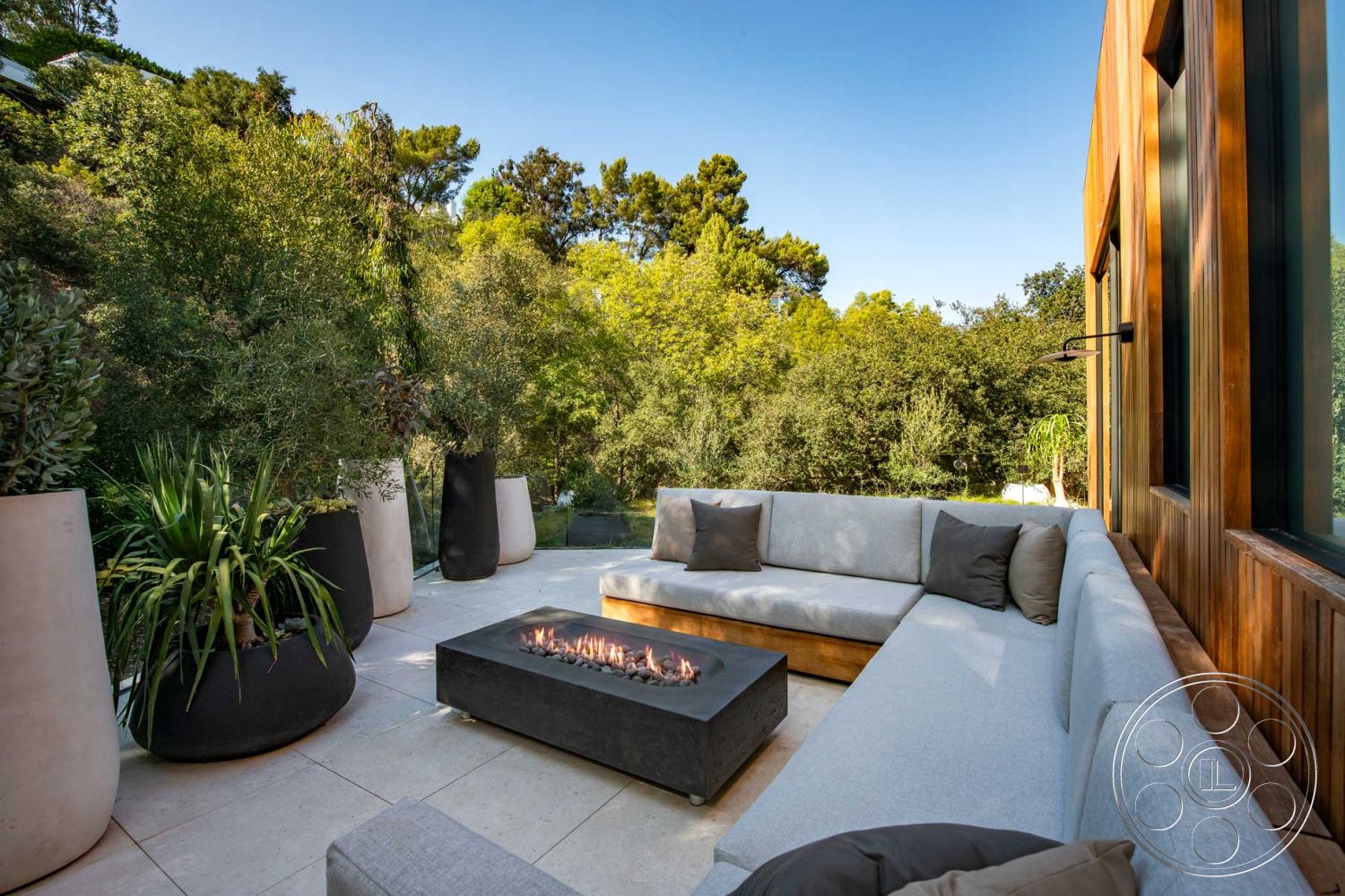 Modern 520 - concrete patio flooring, lush greenery setting, urban garden retreat, floor-to-ceiling glass windows, natural light environment, rectangular fire pit, open-air entertaining space, exterior lounge area, comfortable outdoor cushions, modern outdoor sectional sofa, dark stone fire pit countertop, textured concrete planter pots, surrounding tree landscape, wooden panel accents