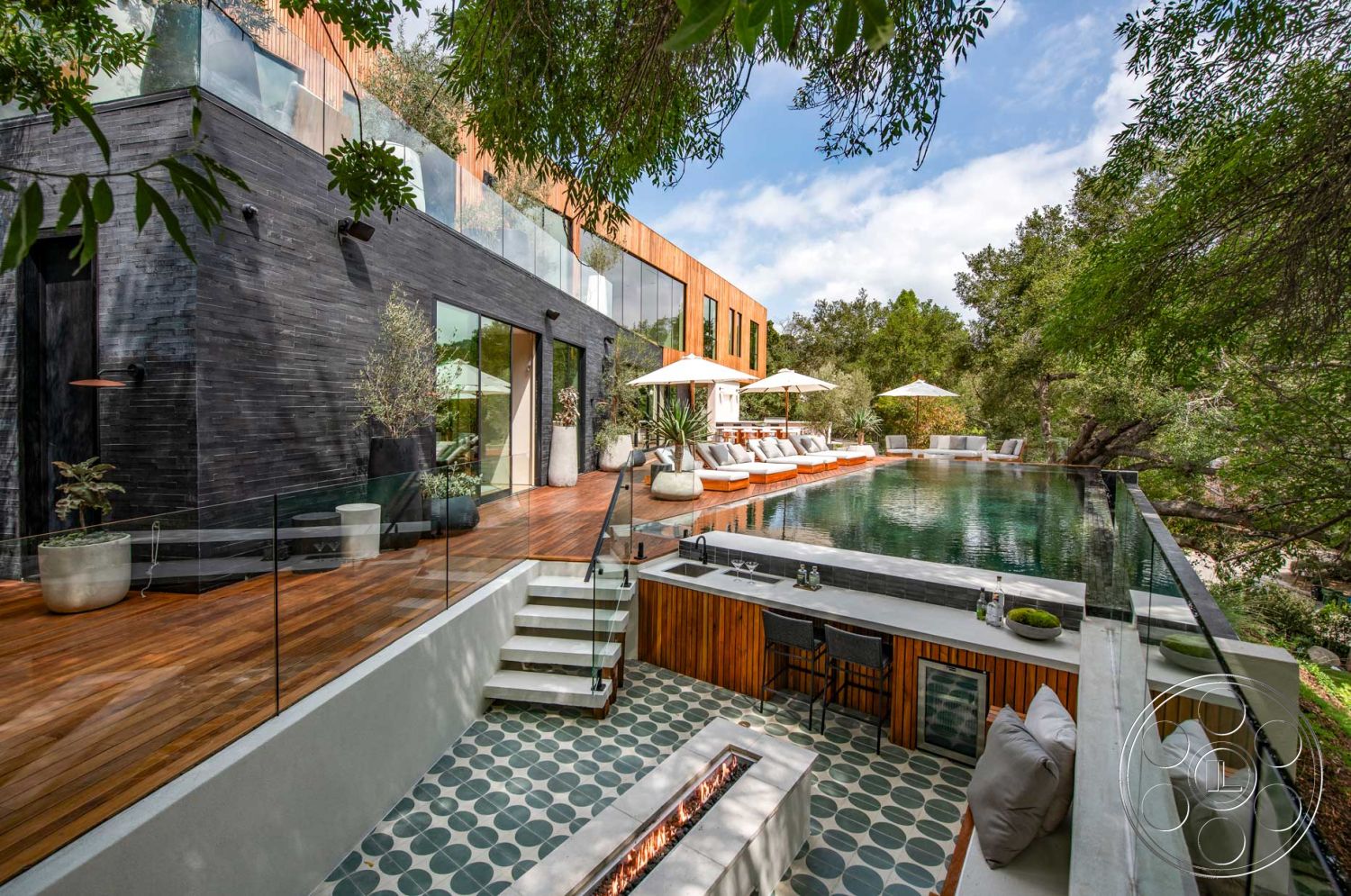 Modern 520 - exterior, nature view, glass railing, infinity pool, open-air living space, surrounded by trees, cozy outdoor seating, floor-to-ceiling glass windows, wood deck patio, geometric patterned tile flooring, rectangle infinity pool, contemporary lounge chairs, stone fire pit seating area, polished concrete outdoor kitchen, black stone feature wall, modern wooden slat fencing, umbrella-shaded patio area, wooden privacy screens