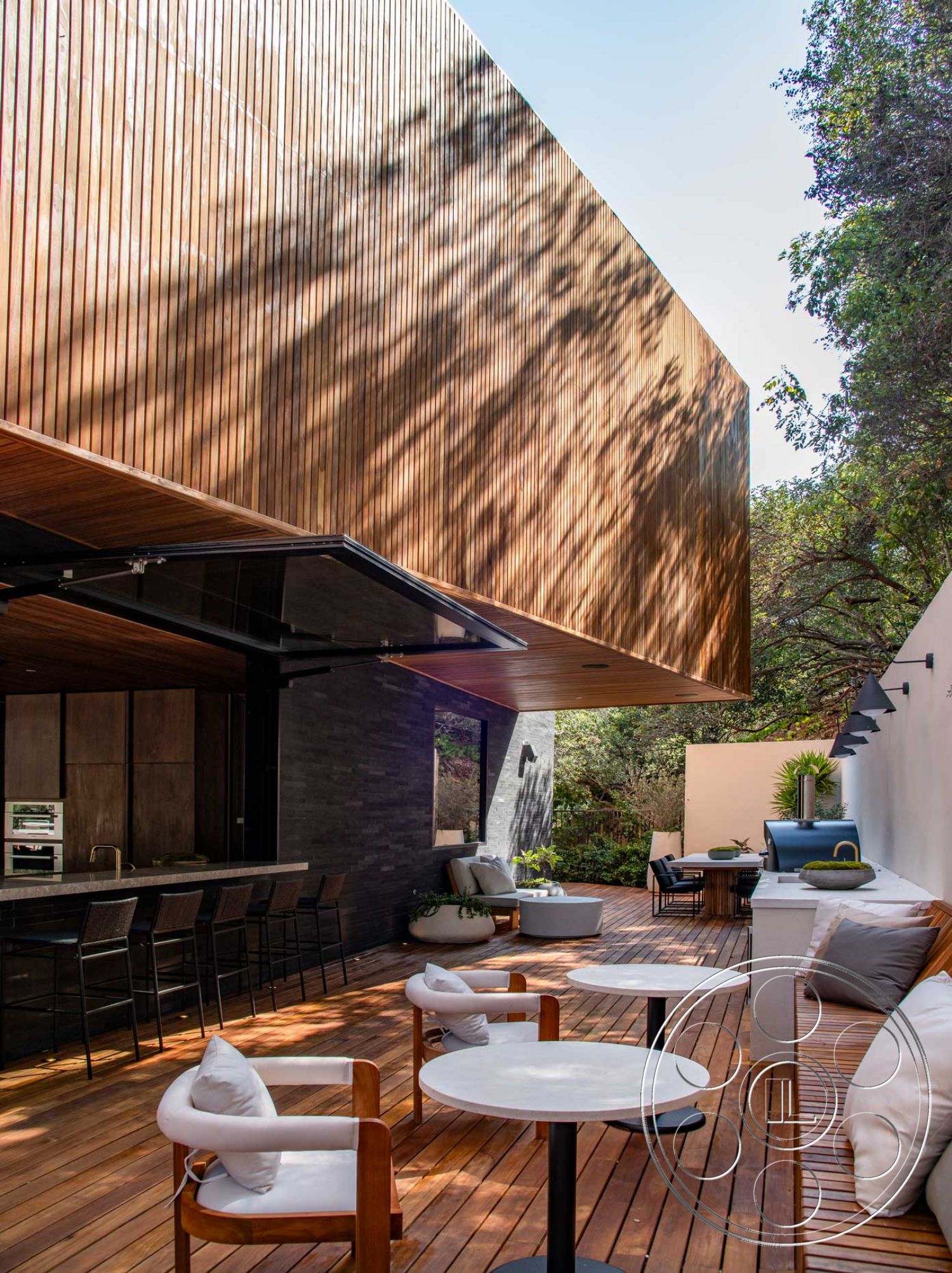 Modern 520 - metal bar stools, open-air living space, contemporary architectural style, floor-to-ceiling kitchen windows, surrounding lush greenery, wood deck flooring, minimalist outdoor furniture, modern outdoor dining area, curved sectional seating, exterior wood slat siding, exterior stone accent wall, outdoor kitchen with granite countertop, shadowed light patterns, wooden pergola overhead, planted landscaping features