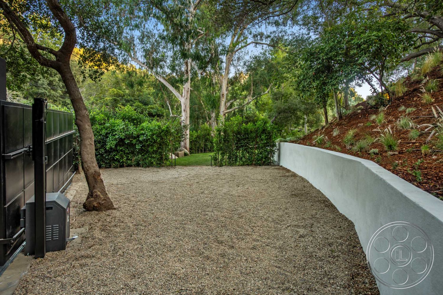 Modern 520 - lush greenery, modern landscape design, landscaped hillside, gravel driveway, concrete retaining wall, sloped garden terrain, natural wood fence, exterior wall with white stucco finish, mixed foliage planting bed, outdoor space with surrounding trees, gate access for driveway, rural property setting, sun-dappled area, interior view of exterior features