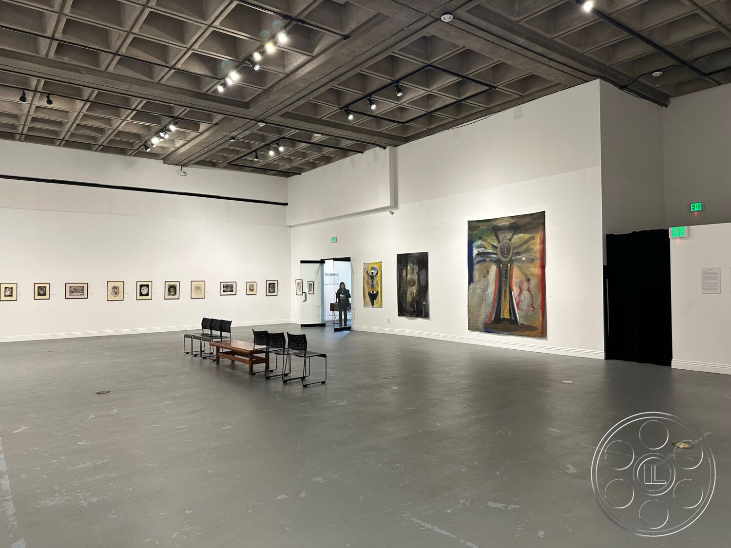 Art Gallery DTLA - track lighting fixtures, exposed concrete ceiling panels, gray polished concrete floor, crisp architectural lines, white painted gallery walls, minimalist art gallery interior, modernist open floor plan, framed artwork installations, black curtain partition, rectangular seating area with wooden coffee table, two-tone color scheme with white and gray, exterior entrance with glass door, spacious exhibition space, urban cultural setting