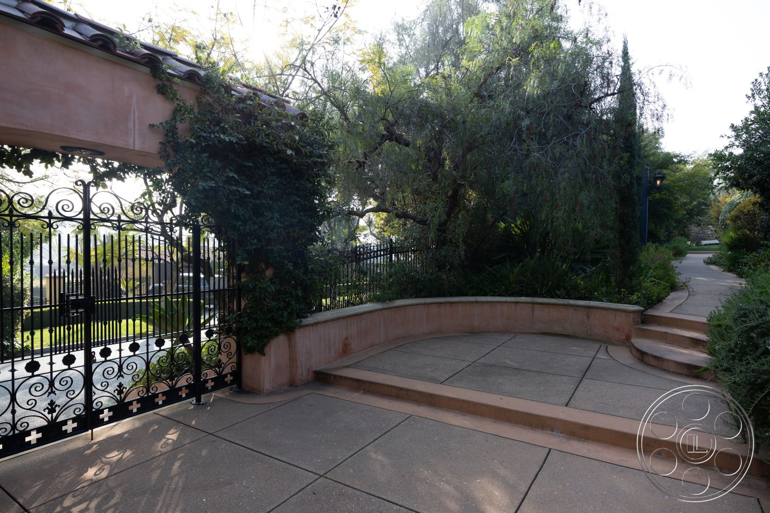 Mansion 145 - paved driveway, fenced garden area, textured concrete patio, stucco walls, arched entryway, architectural detail, decorative wrought iron, integrated landscaping, natural stone retaining wall, flagstone pathway, mature tree canopy, exposed aggregate concrete, lush garden landscaping, curved concrete steps, urban outdoor setting, wooden support beams, iron gate entrance, sandy-colored stucco, garden pathway lighting, rounded patio edge