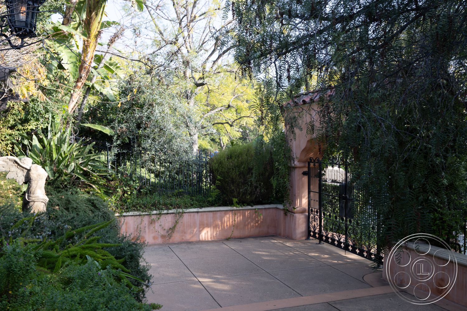 Mansion 145 - exterior, green shrubbery, lush tropical landscaping, planting beds, textured concrete surface, wrought iron gate, concrete patio flooring, curved garden path, sunlit outdoor area, private garden retreat, stone garden statue, pink stucco garden walls, mature trees setting, border plantings, ornamental iron fence