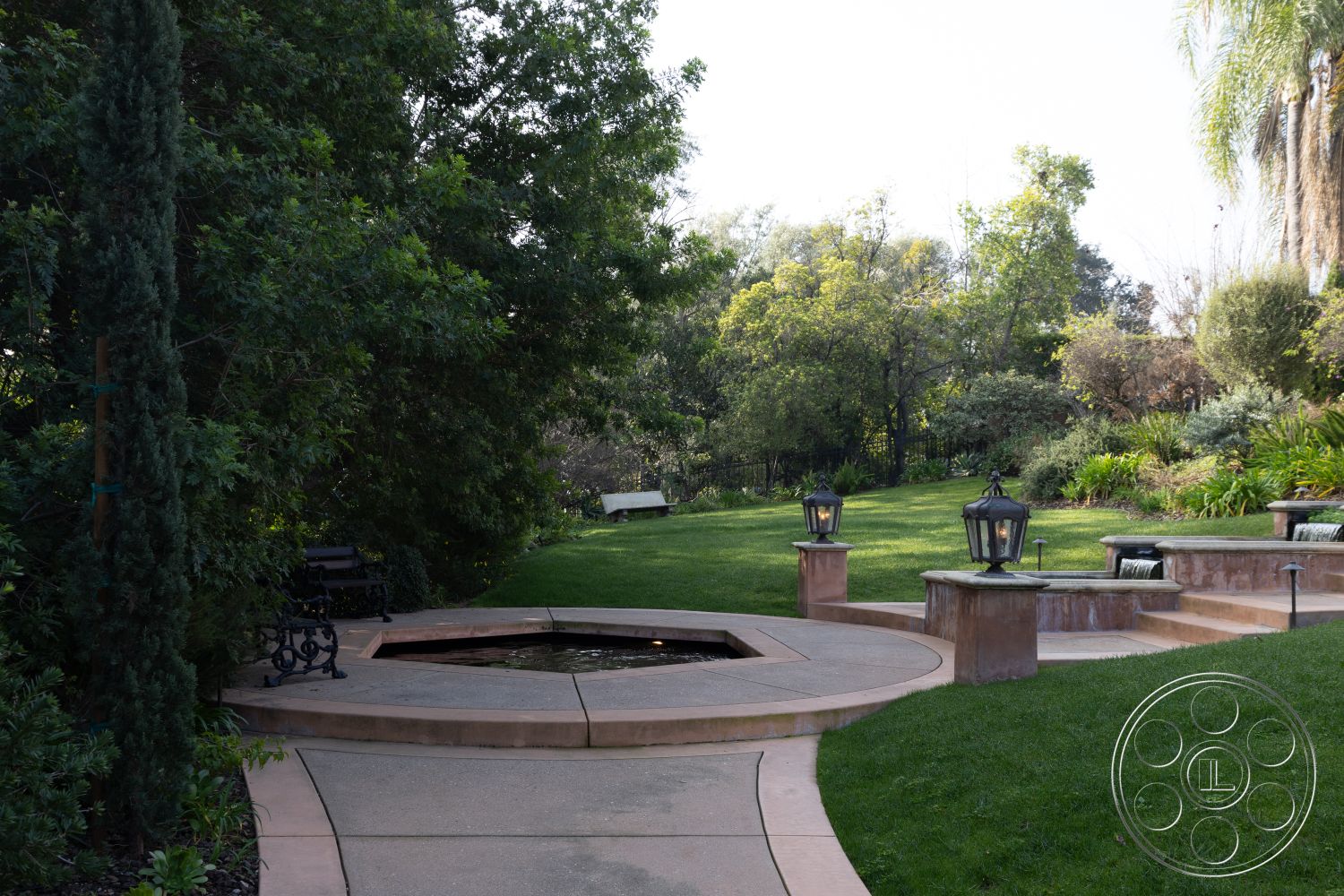 Mansion 145 - lush green lawn, landscaped garden area, exterior landscape design, exposed aggregate patio surface, decorative outdoor lanterns, cypress trees lining the path, paved stone walkways, circular concrete patio, freestanding garden fountain, stepped stone waterfall feature, benches in garden setting, multi-tiered garden beds, natural vegetation backdrop, tranquil outdoor ambiance, serene private garden retreat, sunny garden location, botanical landscape, peaceful garden oasis, relaxation space in nature