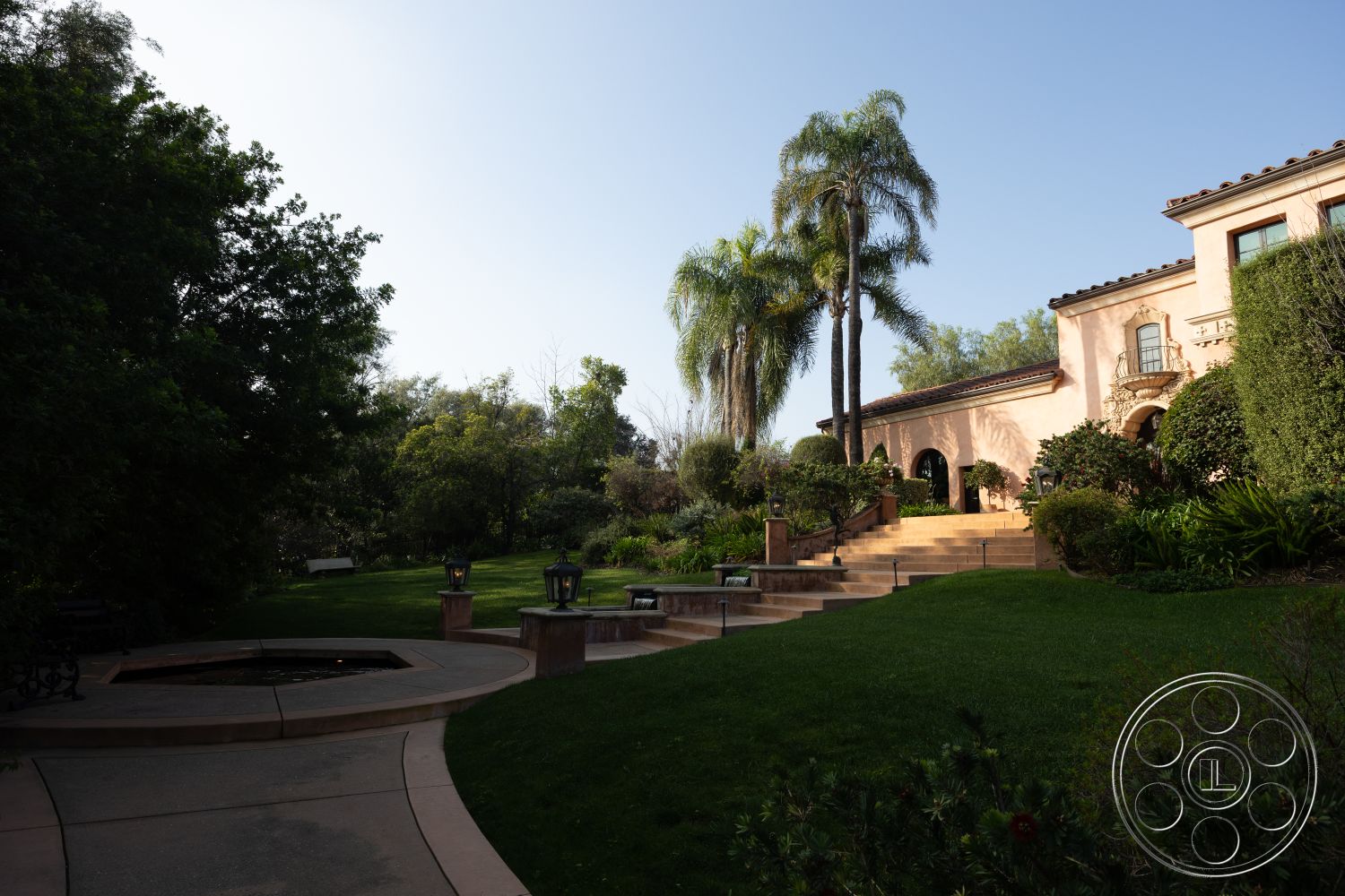 Mansion 145 - exterior, outdoor seating area, terracotta roof tiles, arched doorways, formal garden, ornamental bushes, well-maintained yard, Mediterranean architectural style, elevated patio, two-story home, wrought iron railings, lush greenery surrounding, curved stone pathway, stucco house facade, landscaped garden with palm trees, sloped lawn area, decorative stone steps, outdoor lantern light fixtures, stone fountain feature, pathway with concrete border