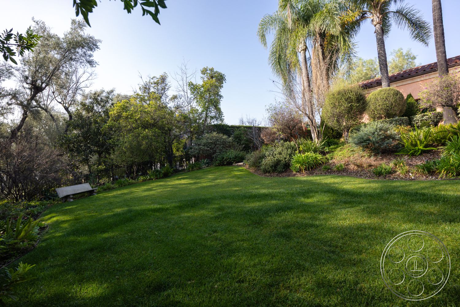 Mansion 145 - landscaped yard, lush green lawn, surrounding foliage, tropical palm trees, suburban backyard, sunny outdoor space, planted flower beds, exterior garden setting, natural light environment, natural stone edging, bench seating area, varied shrubbery, well-maintained grass, soft shadows from trees, inviting relaxation spot