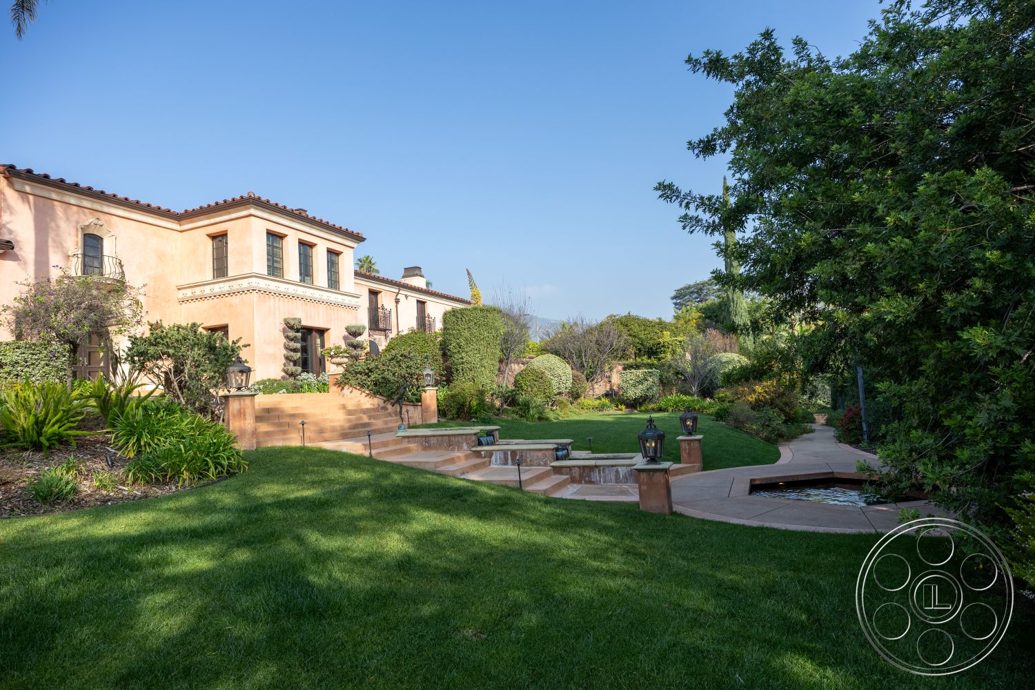 Mansion 145 - exterior, modern water feature, vibrant flower beds, arched doorways, tile roof, Mediterranean architectural style, stone bench, stucco exterior walls, urban garden setting, cobblestone patio area, landscaped garden with tropical plants, grassy lawn area, decorative wrought iron balcony railings, multiple outdoor seating areas, circular stone garden pathway, hardscape steps leading to the entrance, outdoor lantern lighting