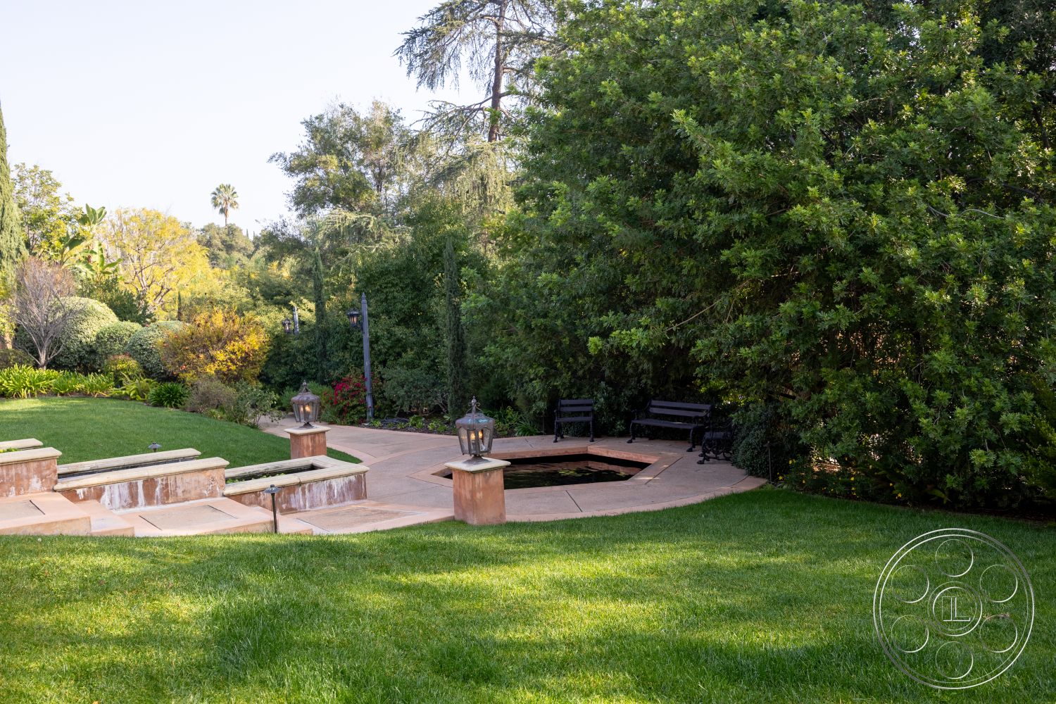 Mansion 145 - exterior garden patio, lush greenery surrounding outdoor space, concrete patio floor, curved garden pathways, decorative outdoor lanterns, lawn area with manicured grass, stone fire pit seating area, wooden benches in garden setting, shrubbery landscaping with flowering plants, natural shaded canopy from trees, benches near fire pit area, tranquil suburban backyard environment, open layout with inviting outdoor seating