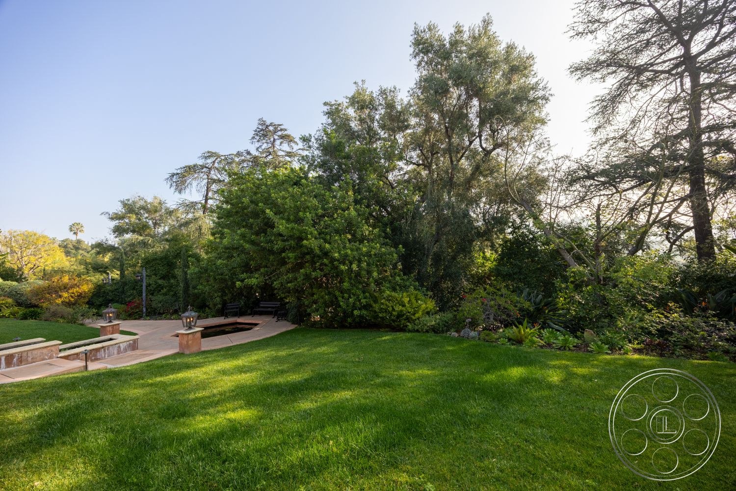 Mansion 145 - natural stone accents, lush greenery, wooden bench, concrete pathway, landscaped lawn, park-like setting, outdoor lighting fixtures, elevated landscaping, exterior garden patio, open sky view, curved garden paths, brick seating area, ornamental flower beds, trees surrounding property, tranquil backyard environment, peaceful retreat space