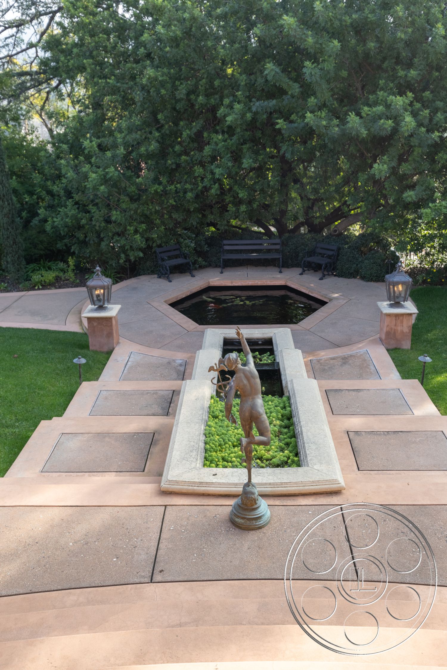 Mansion 145 - exterior, manicured lawn, landscape greenery, outdoor relaxation space, concrete patio flooring, surrounding trees and shrubs, decorative lantern light fixtures, curved concrete walkways, garden patio setting, symmetrical landscaping design, octagonal water feature, sculpted garden statue, black metal bench seating, framed flowerbed with greenery, tranquil garden atmosphere