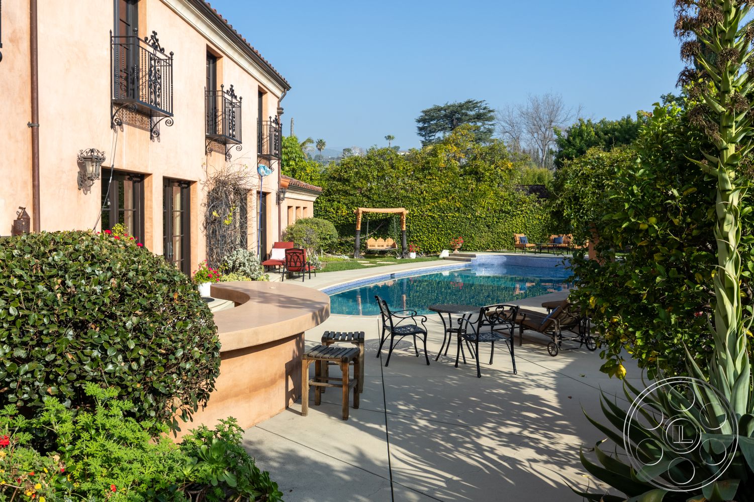 Mansion 145 - exterior, landscaped garden, fenced garden area, tropical landscaping, built-in seating, metal balcony railings, concrete pool deck, rectangle swimming pool, stucco exterior walls, lush greenery surrounding, sunny backyard setting, brick outdoor seating area, curved outdoor bar, wooden gazebo structure, painted window frames, textured patio surface, decorative outdoor light fixtures, outdoor dining furniture, wood and wrought iron furniture