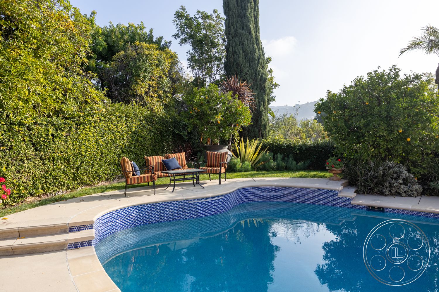 Mansion 145 - outdoor seating area, rectangular swimming pool, metal coffee table, exterior concrete pool border, lush green foliage, relaxing garden atmosphere, concrete pool steps, tranquil outdoor environment, blue mosaic tile pool edge, grass landscaping around pool, wooden patio chairs with cushions, tropical plants in garden, bushes and hedges surrounding pool, sunny hillside setting, mountain view in background