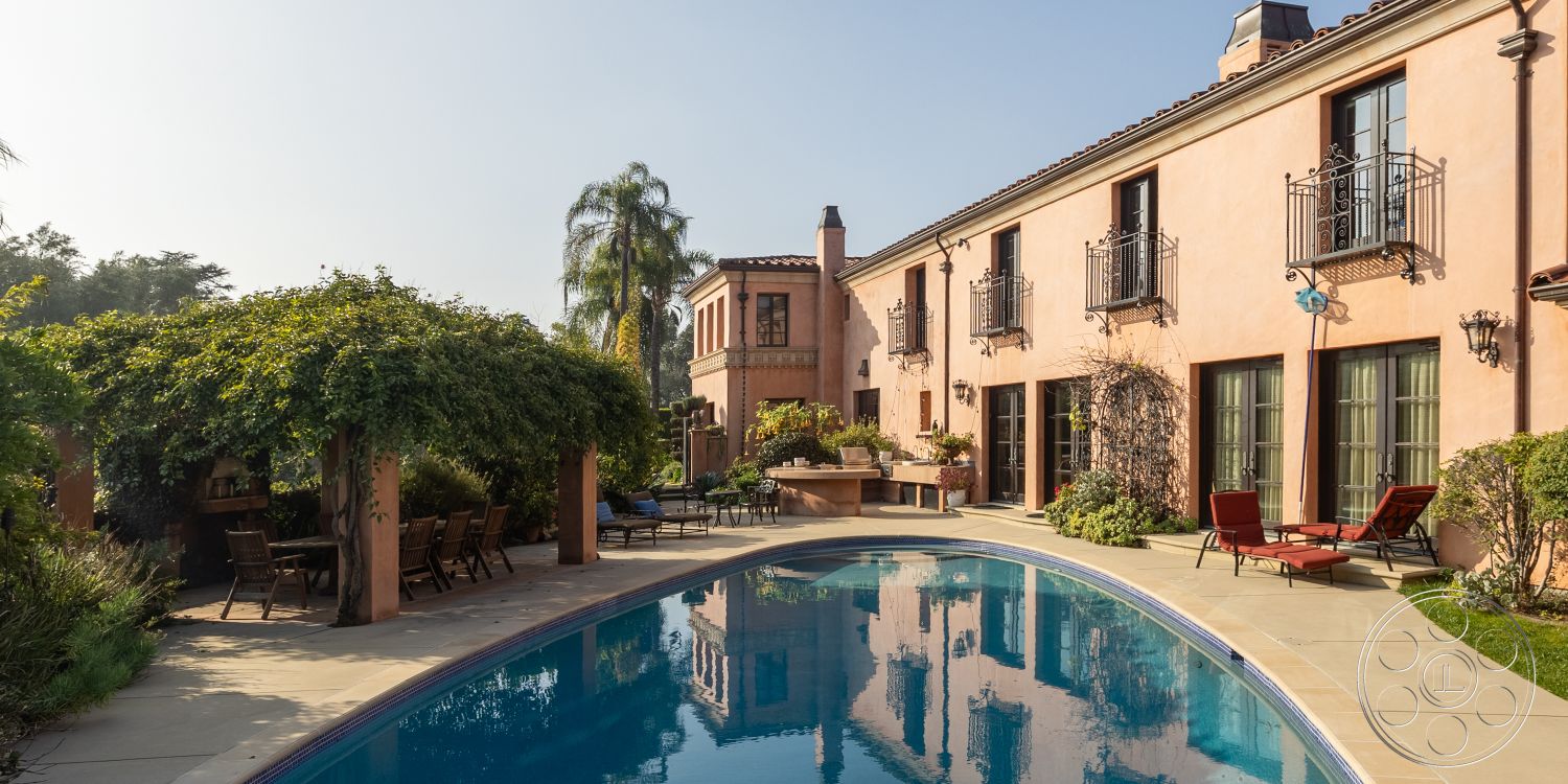 Mansion 145 - exterior, landscaped garden, rectangular swimming pool, outdoor dining set, sunny courtyard, arched windows, Mediterranean architectural style, textured stucco walls, luxury outdoor seating, palm tree landscaping, floor-to-ceiling glass doors, indoor-outdoor living space, concrete pool decking, iron balcony railings, wood pergola with climbing vines, terracotta tiled roof, backyard pool area