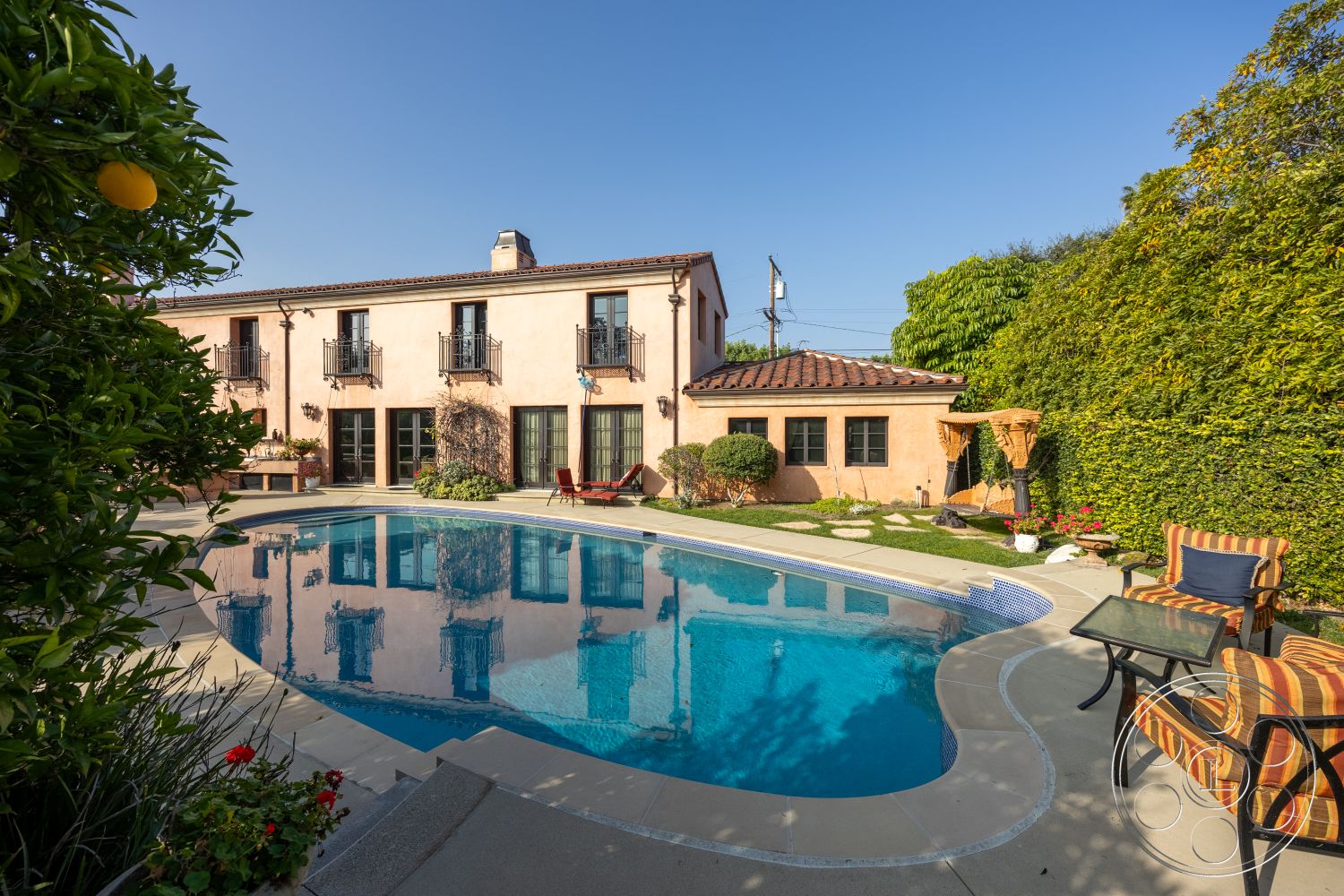 Mansion 145 - exterior, rectangular swimming pool, floor-to-ceiling windows, outdoor seating, relaxation area, landscaped backyard, terracotta roof tiles, arched doorways, concrete pool deck, wooden patio furniture, sunny outdoor setting, mediterranean architectural style, decorative stone features, lush green backdrop, plastered stucco walls, stone pool coping, garden with hedges, black wrought iron balcony railings, paved outdoor patio, surrounding citrus trees