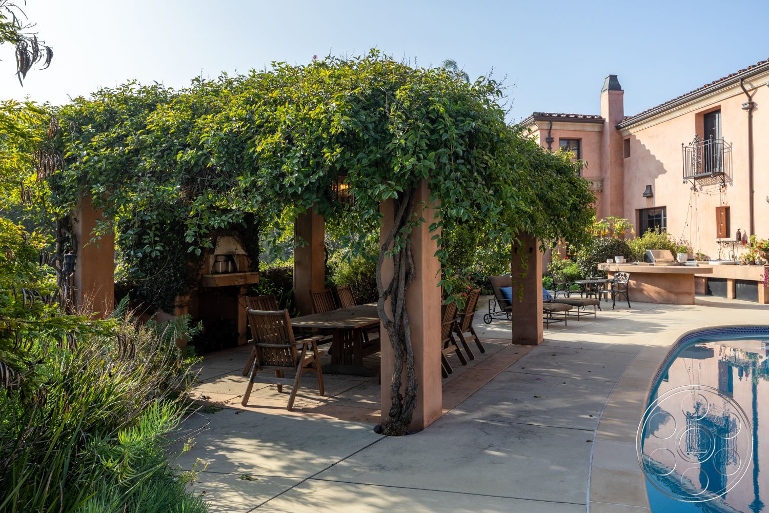 Mansion 145 - wooden dining table, rectangular swimming pool, outdoor dining area, spacious backyard, Mediterranean architectural style, poolside lounge area, sunny outdoor setting, landscaped garden area, concrete patio flooring, stucco exterior walls, exposed wood beam pergola, wooden patio chairs, brick outdoor fireplace, climbing vines on structure, potted plants surrounding space