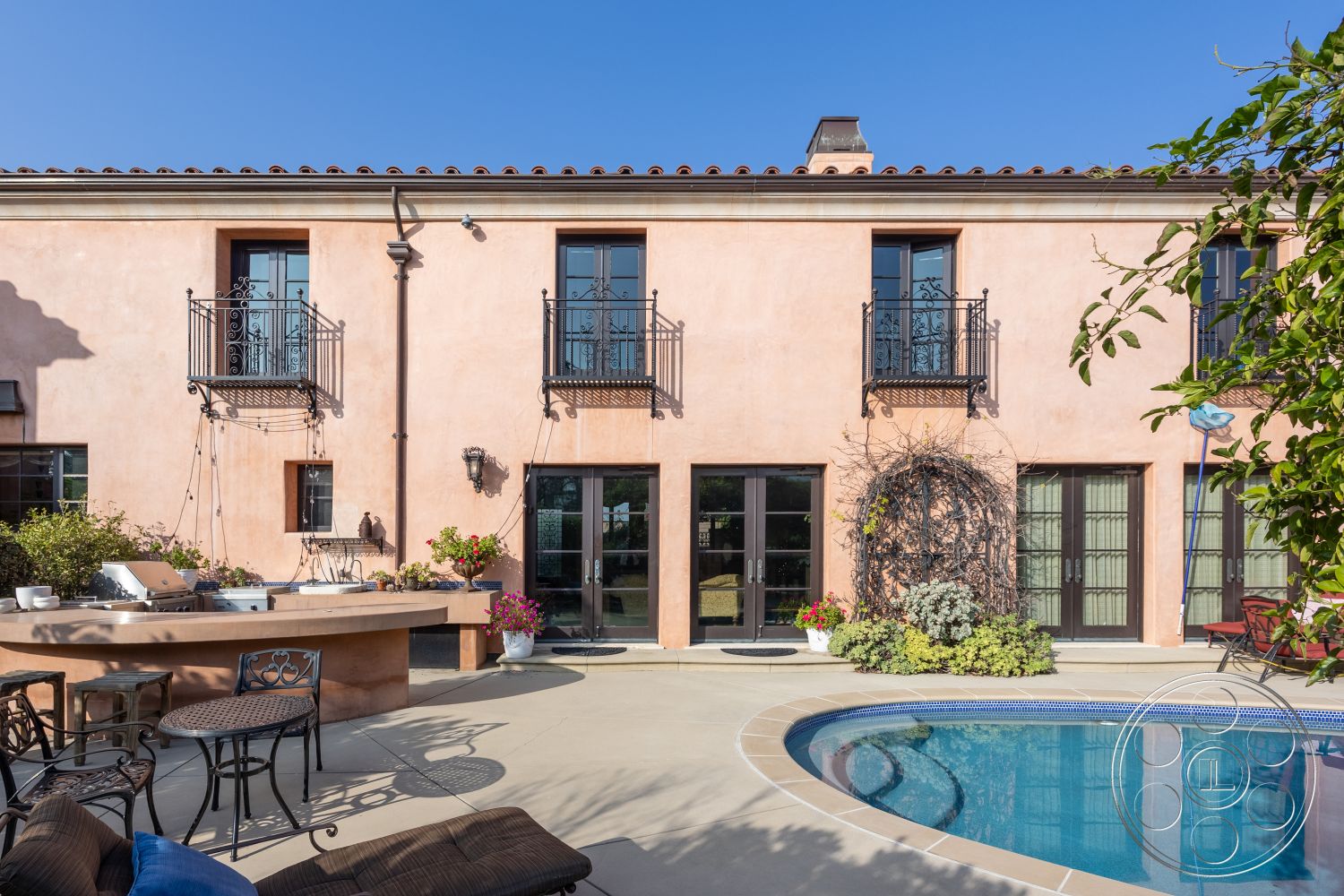 Mansion 145 - rectangular swimming pool, Mediterranean architectural style, concrete patio flooring, exterior stucco walls, arched windows with black frames, exposed concrete pool border, plastered wall finish, black wrought iron balcony railings, tile roofing with terracotta tiles, stone outdoor kitchen countertop, wooden outdoor dining chairs, green landscaping with flowering plants, double glass doors leading to the pool area, court-yard style outdoor space, sunny urban backyard setting, pre-cast concrete pool deck, decorative outdoor lighting fixtures