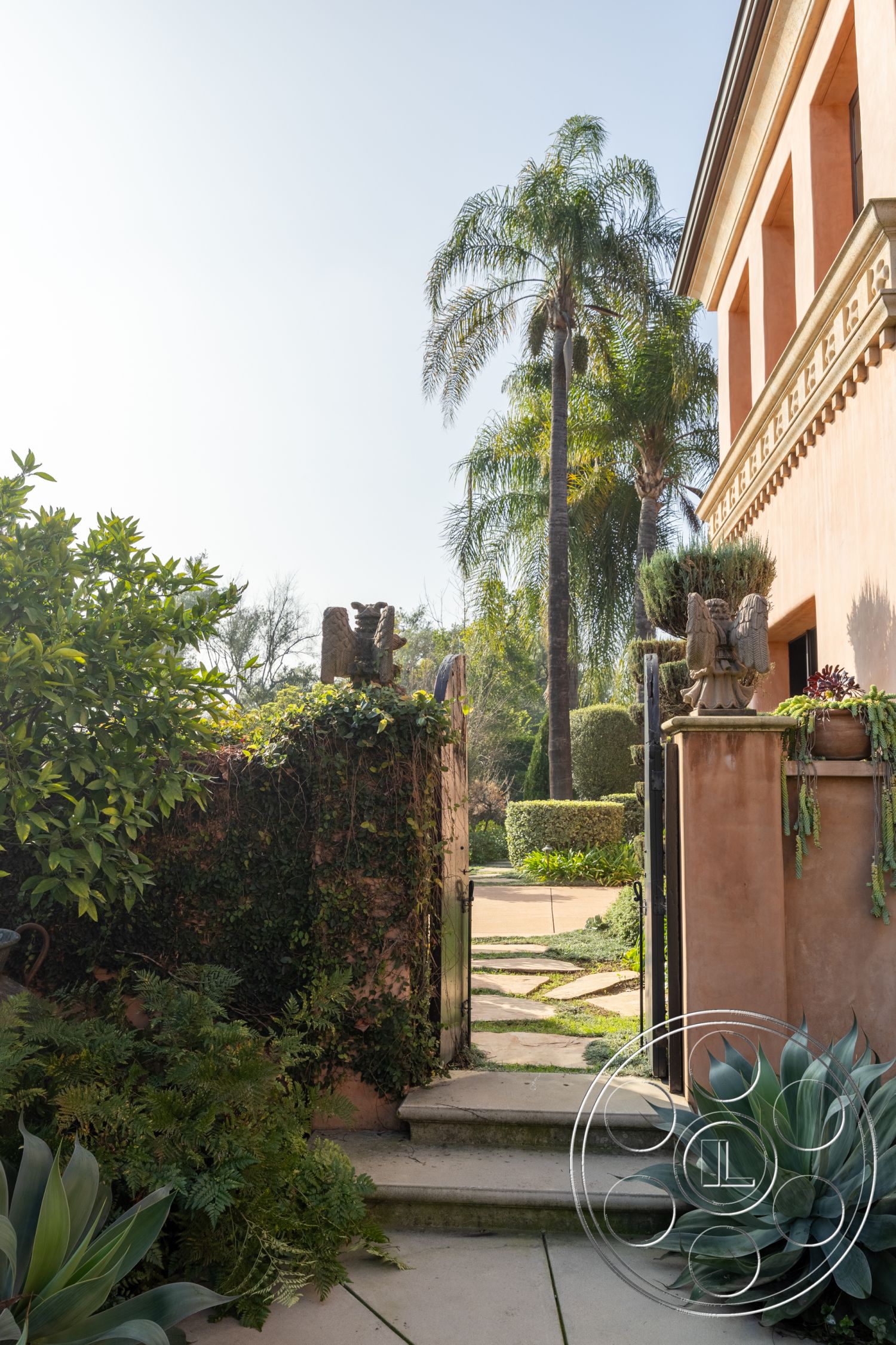 Mansion 145 - natural light, landscaped garden, palm trees, stone stepping stones, vibrant greenery, textured plaster walls, garden entrance, exterior garden pathway, lush tropical plants, decorative wrought iron gate, modern outdoor aesthetic, rustic element with ivy, softly curved garden beds