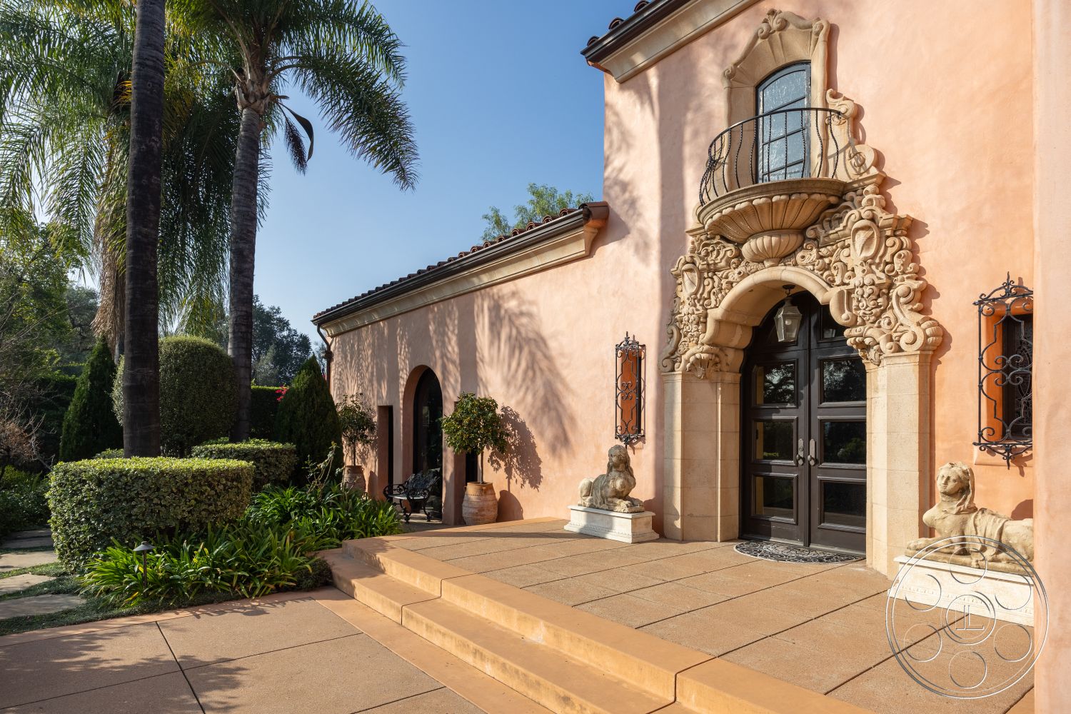 Mansion 145 - exterior, terracotta roof tiles, iron balcony railing, Mediterranean architectural style, stucco wall finish, lush greenery surrounding property, landscaped garden with palm trees, courtyard-style layout, decorative stone entrance arch, ornate plaster molding, circular concrete steps, manicured hedges and shrubs, double front doors with glass panels, ceramic outdoor lanterns, sculptural stone lion statues, formal entryway patio, tiled walkway, arched doorways leading to interior, balcony with decorative ironwork