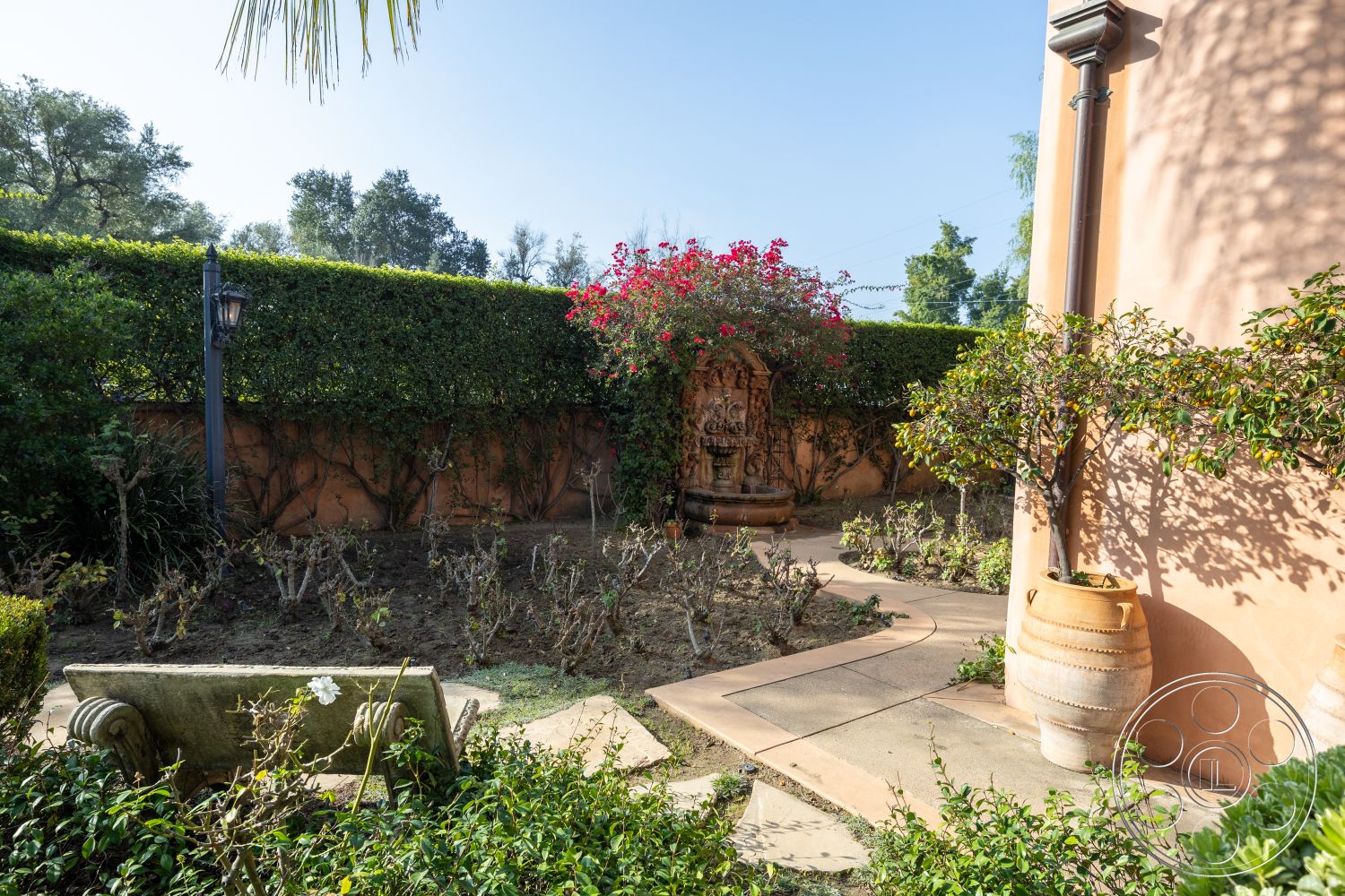 Mansion 145 - exterior garden patio, lush greenery landscaping, exposed aggregate concrete path, curved stone pathway, flowering bougainvillea plants, decorative stone fountain, terracotta garden planters, stucco wall with peach finish, ornamental flowering plants, traditional wrought iron lantern, low stone bench seating, mediterranean garden setting, planted hedges for privacy, terracotta pottery accents, natural light garden area