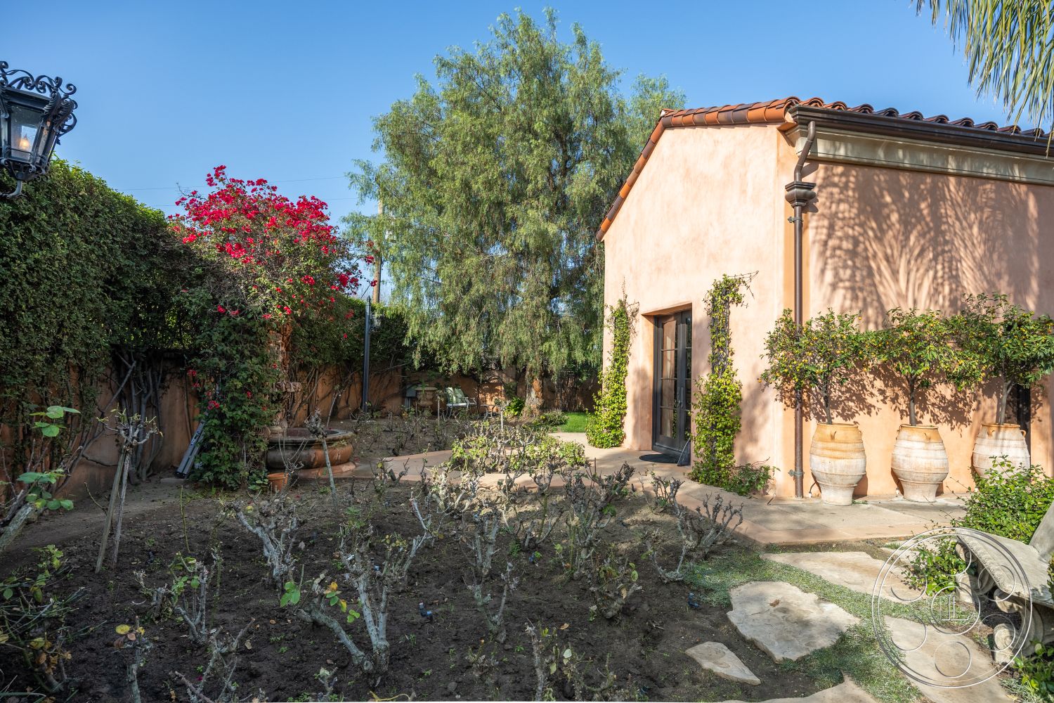 Mansion 145 - mature trees, stone pathway, secluded backyard, terra cotta roof tiles, paved courtyard, Mediterranean architectural style, flowering bougainvillea, exterior stucco walls, lush garden setting, landscaped garden beds, wooden garden bench, vintage lantern light fixture, rustic clay pots, textured plaster wall surface, natural stone hardscaping