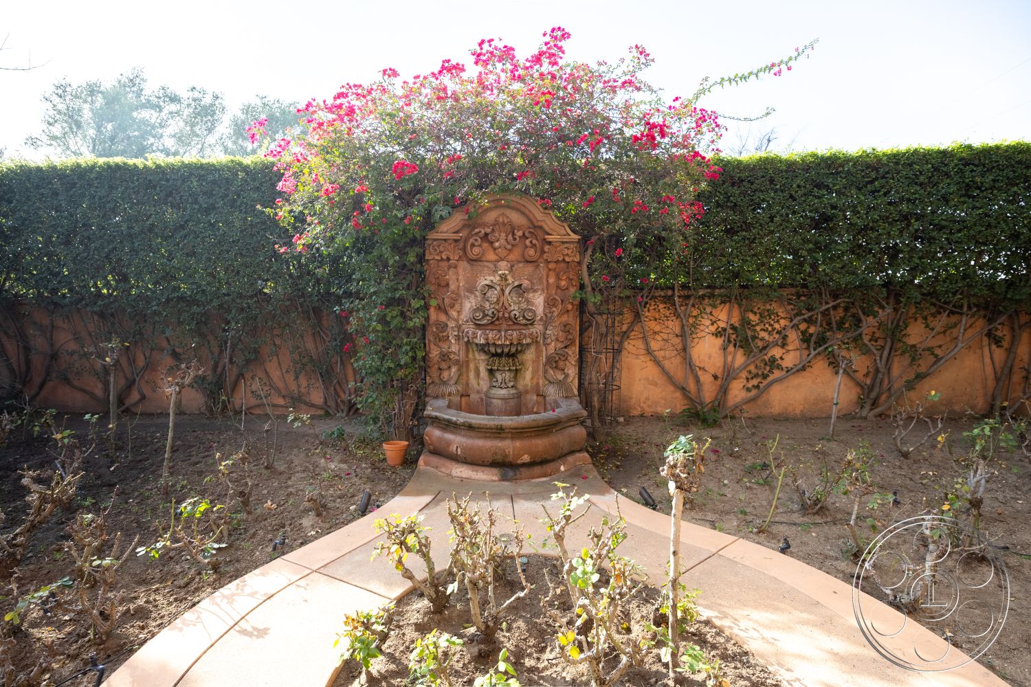 Mansion 145 - landscaped flower beds, exterior garden, sunlit garden setting, surrounding hedges, handcrafted stone fountain, intricate plaster fountain design, vibrant bougainvillea foliage, curved pathway garden design, textured adobe wall, earthy terracotta tones, mediterranean garden style, organic garden layout