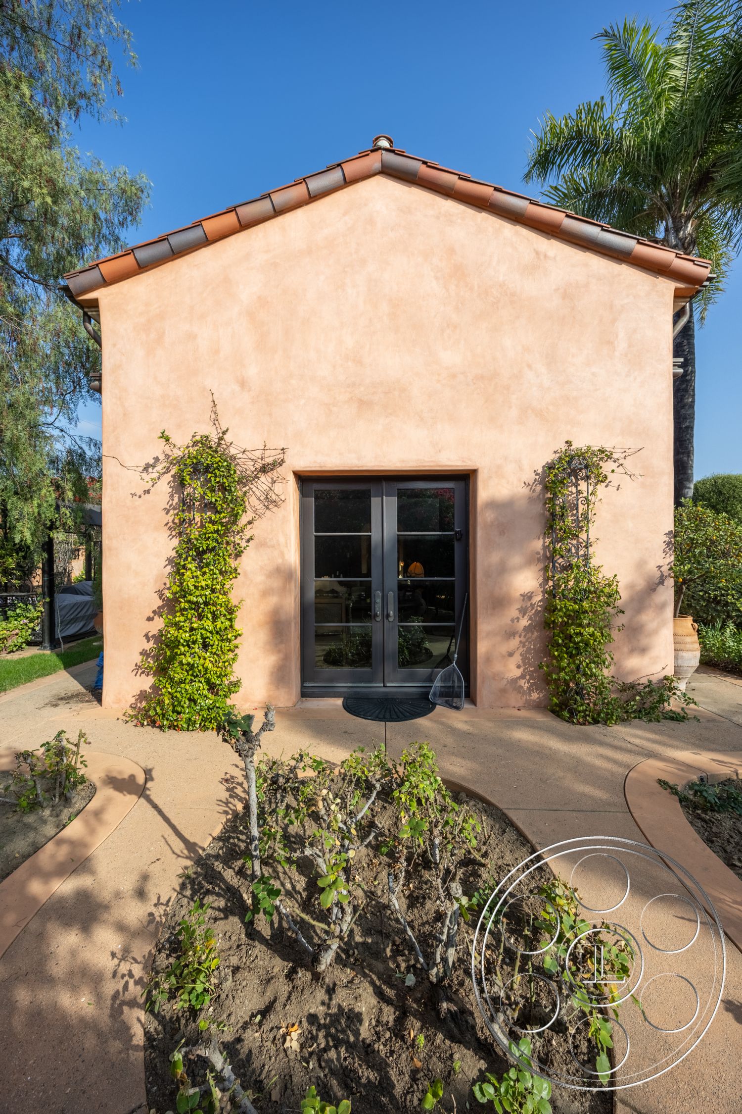 Mansion 145 - modern Mediterranean architecture, exterior stucco wall, landscaped garden beds, terra cotta tile roof, desert garden setting, paved entrance walkway, black double French doors, rounded pathway layout, planted shrubs and foliage, soft ground cover