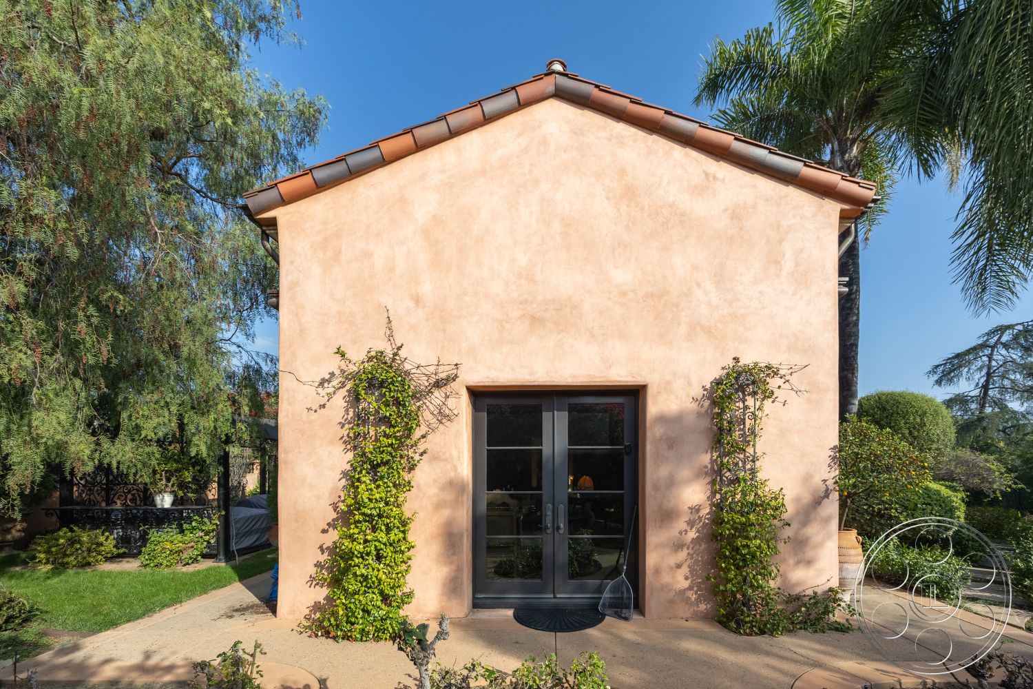 Mansion 145 - exterior, outdoor living space, Spanish tile roof, landscaped garden setting, textured plaster wall, stucco wall finish, flat roof with overhang, paved garden pathway, black steel framed double door, climbing ivy wall treatment, medium-sized leafy trees, lush greenery surrounding structure, traditional Mediterranean style home, arched roofline shape, circular pathway design