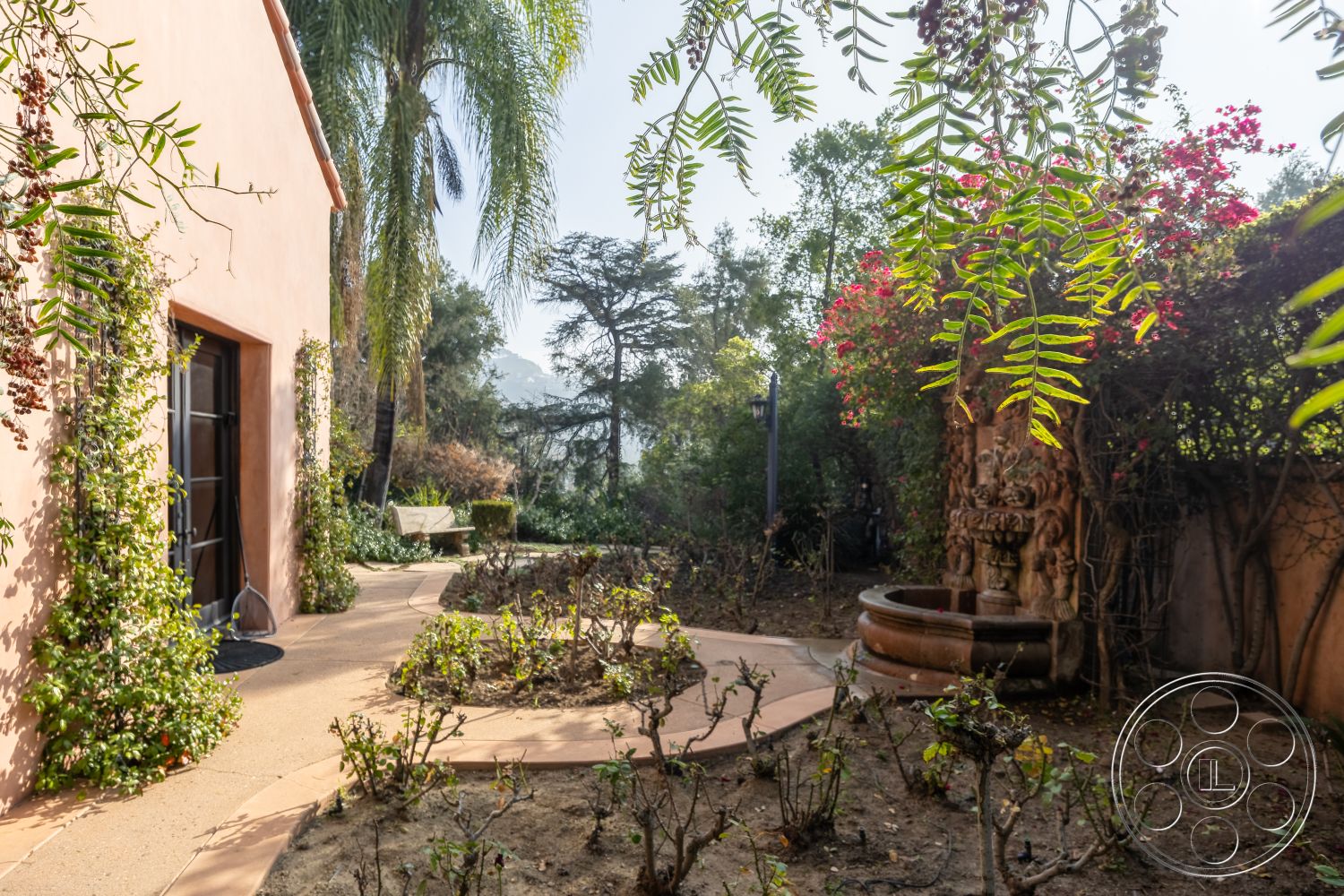 Mansion 145 - landscaped backyard, soft natural lighting, tropical palm trees, sunny outdoor space, exterior stucco walls, lush garden setting, wooden garden bench, curved stone pathway, decorative stone fountain, terracotta garden flooring, walls with climbing vines, flowering shrubs around pathway, rustic outdoor fountain, vibrant garden flowers, view of distant mountains